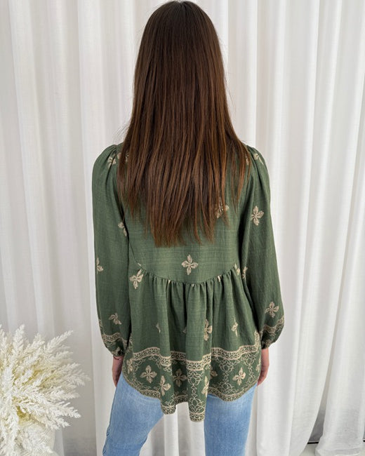 Person wearing a green patterned blouse with long sleeves in front of white curtains