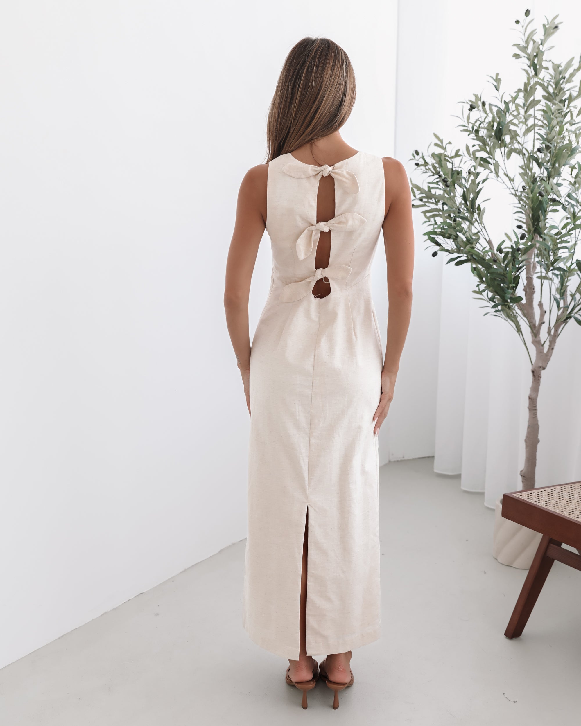 A woman wearing a beige maxi dress with a plunge neckline, bow and slit at the back in a white setting with a wooden chair and a plant
