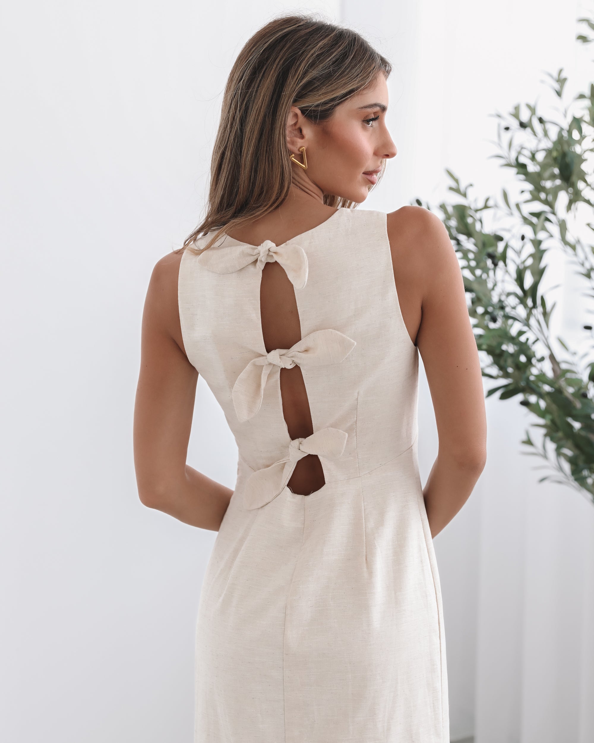 A woman wearing a beige maxi dress with a plunge neckline, bow and slit at the back in a white setting with a wooden chair and a plant
