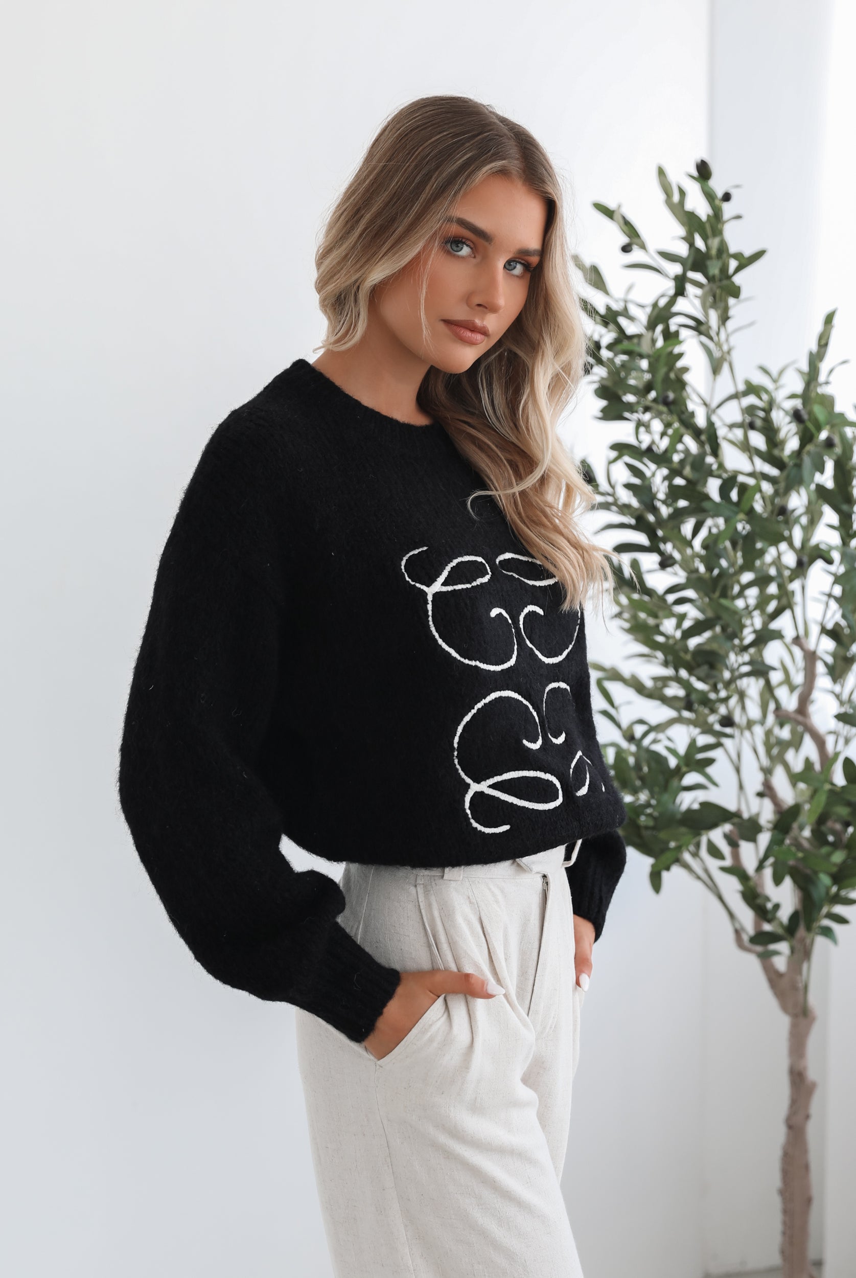 A woman with long, wavy blonde hair is standing indoors near a potted plant, wearing a black sweater with a white abstract design on the front and high-waisted blue jeans. She is looking at the camera with a neutral expression and has one hand resting on her hip. The background is minimalistic with white walls.