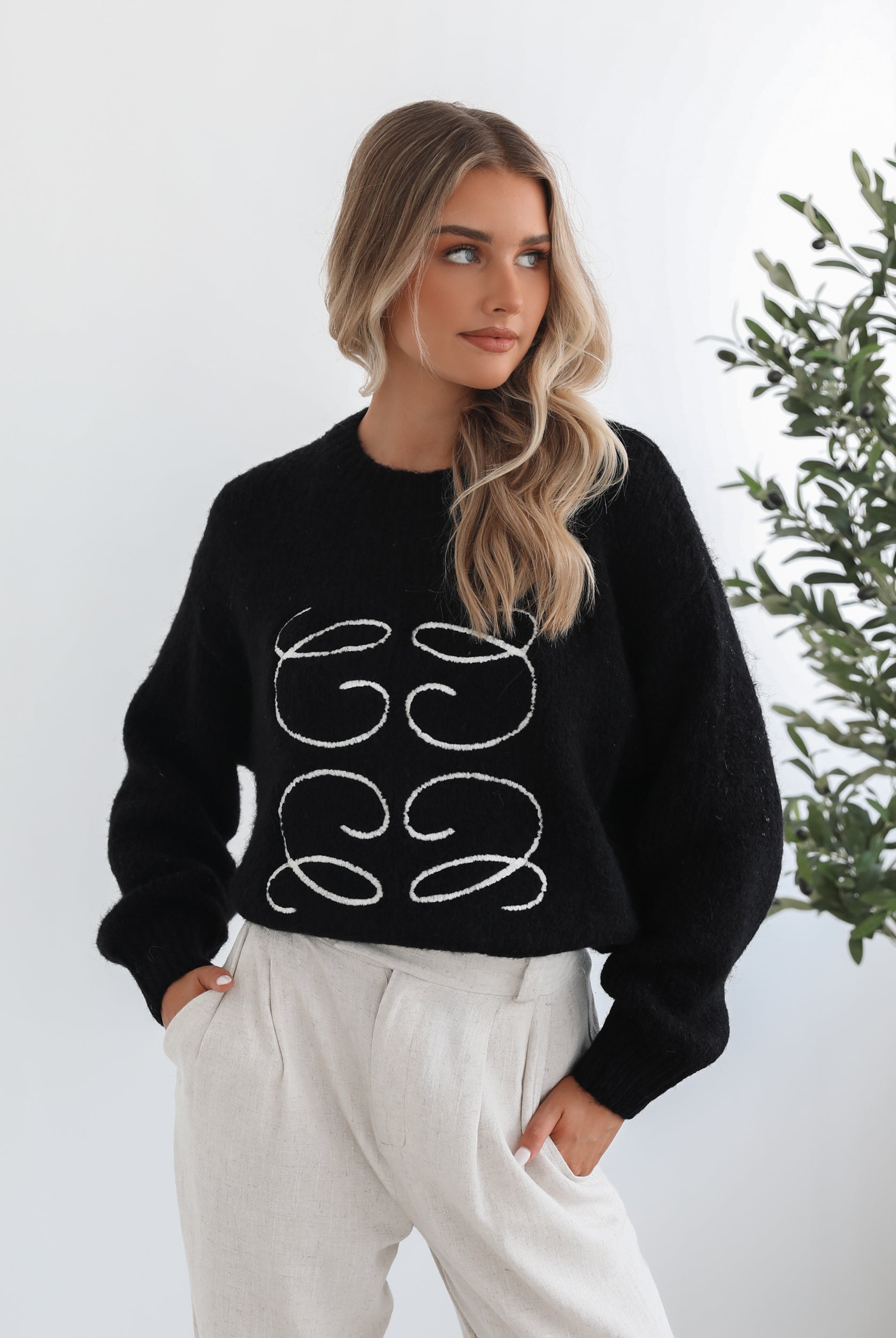 A woman with long, wavy blonde hair is standing indoors near a potted plant, wearing a black sweater with a white abstract design on the front and high-waisted blue jeans. She is looking at the camera with a neutral expression and has one hand resting on her hip. The background is minimalistic with white walls.