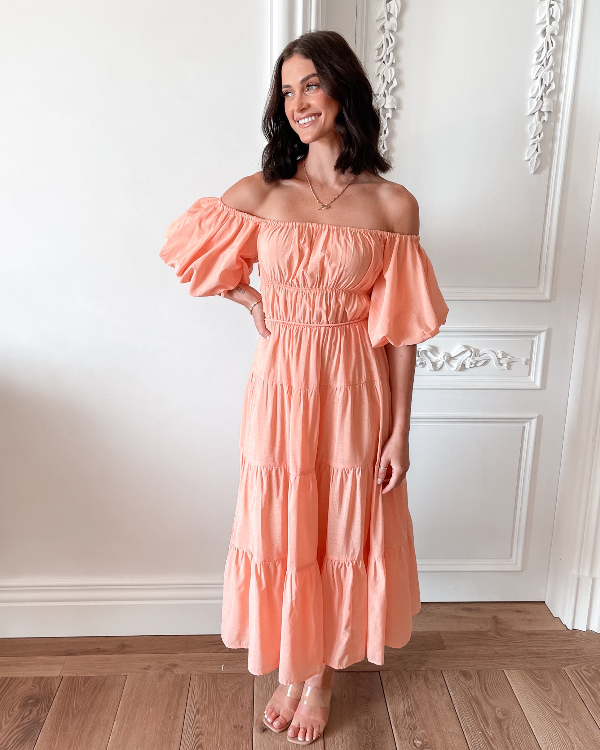 A woman with shoulder-length dark hair stands in a beautifully decorated room with white ornate wall paneling and wooden flooring, wearing an off-the-shoulder peach midi dress with puffed sleeves and a tiered skirt. She is smiling and gently holding one side of her dress, giving a soft and elegant pose.