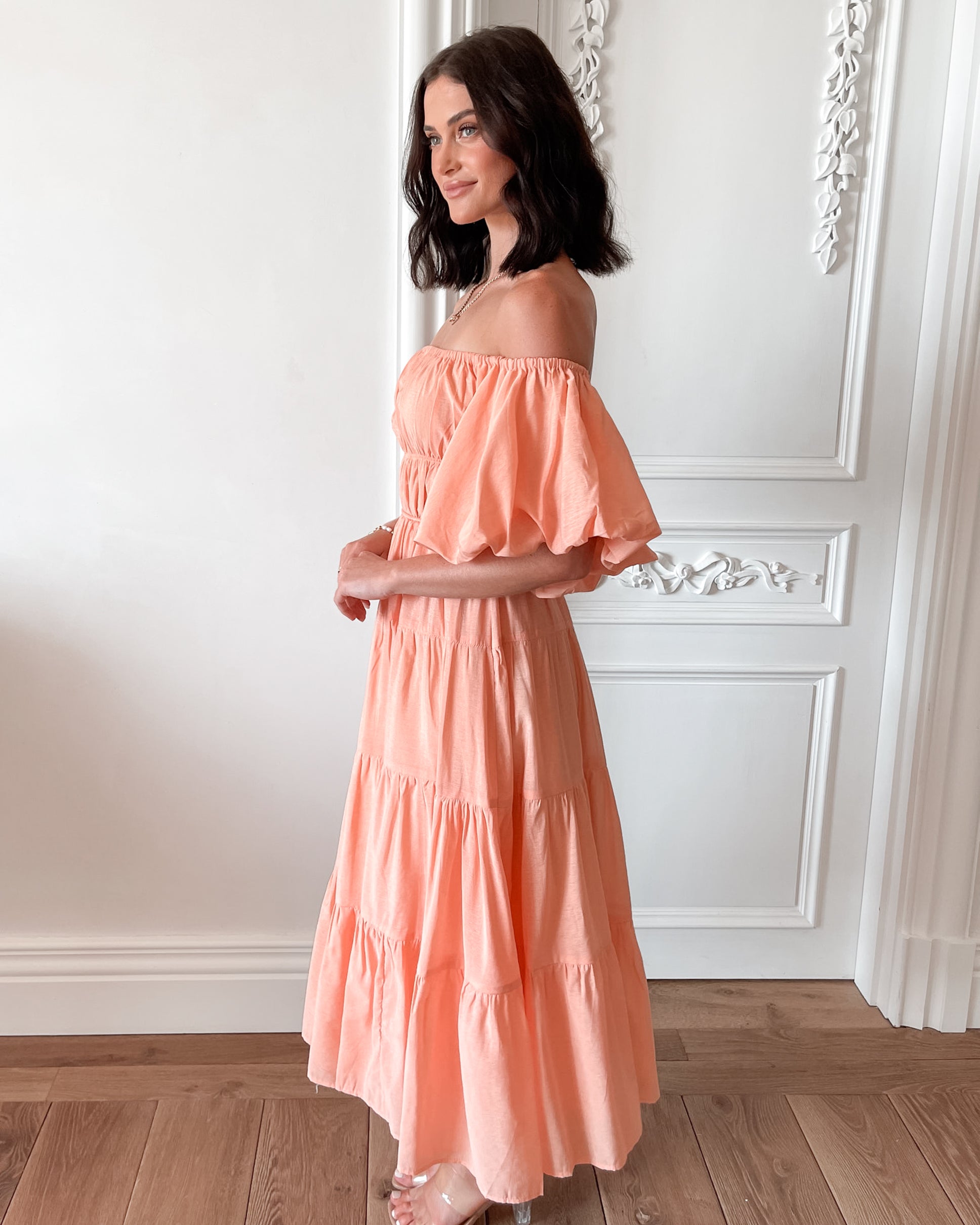 A woman with shoulder-length dark hair stands in a beautifully decorated room with white ornate wall paneling and wooden flooring, wearing an off-the-shoulder peach midi dress with puffed sleeves and a tiered skirt. She is smiling and gently holding one side of her dress, giving a soft and elegant pose.