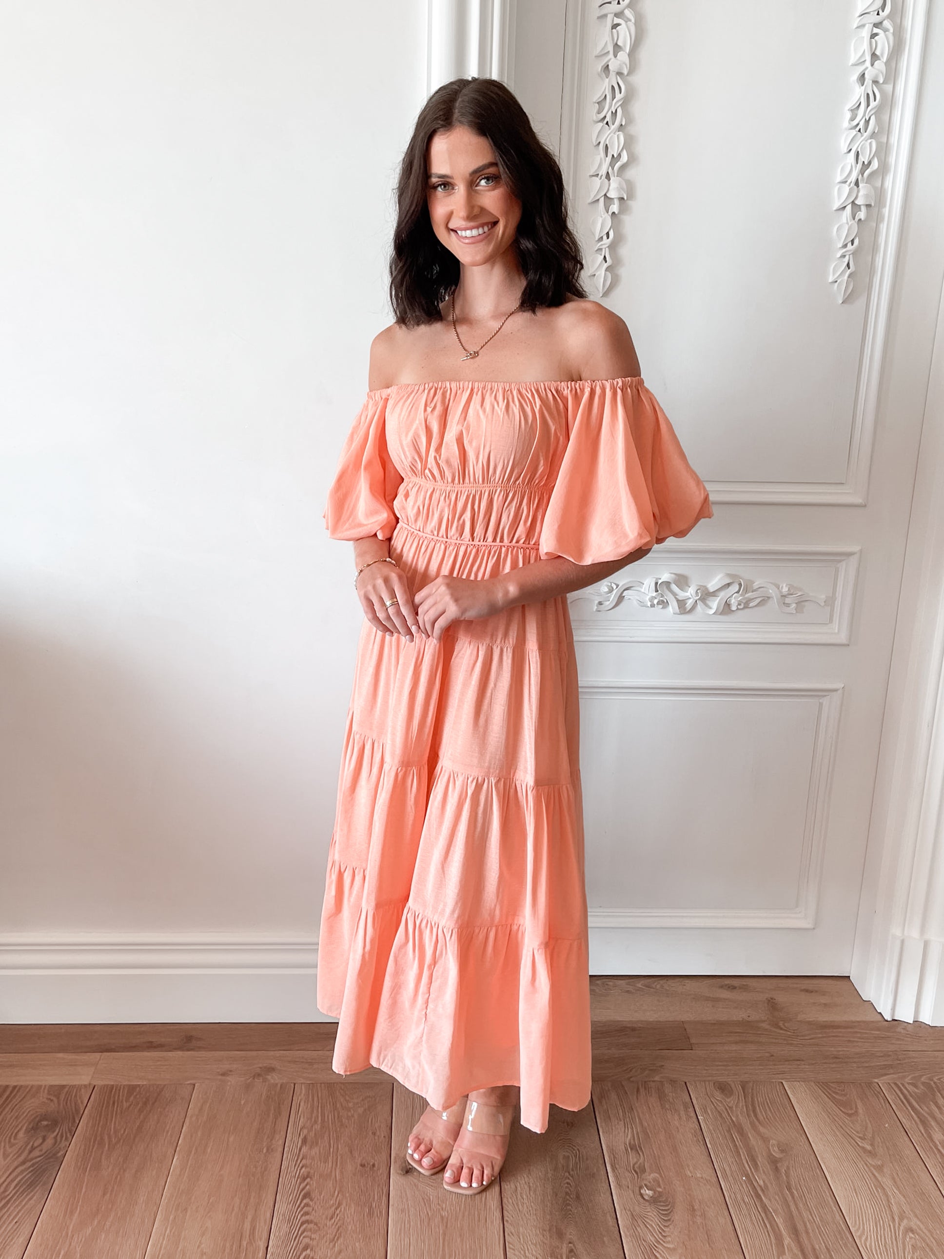 A woman with shoulder-length dark hair stands in a beautifully decorated room with white ornate wall paneling and wooden flooring, wearing an off-the-shoulder peach midi dress with puffed sleeves and a tiered skirt. She is smiling and gently holding one side of her dress, giving a soft and elegant pose.