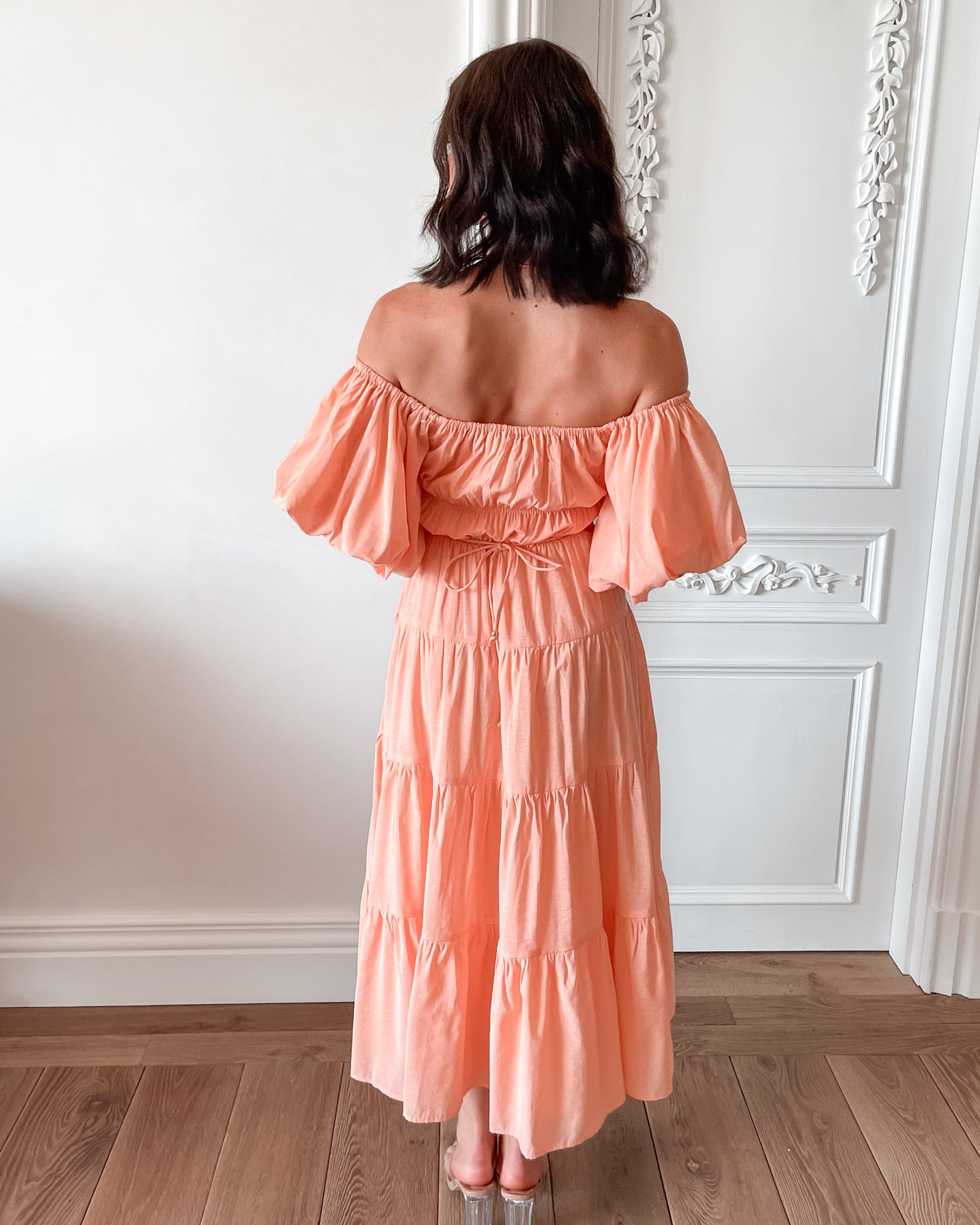 A woman with shoulder-length dark hair stands in a beautifully decorated room with white ornate wall paneling and wooden flooring, wearing an off-the-shoulder peach midi dress with puffed sleeves and a tiered skirt. She is smiling and gently holding one side of her dress, giving a soft and elegant pose.