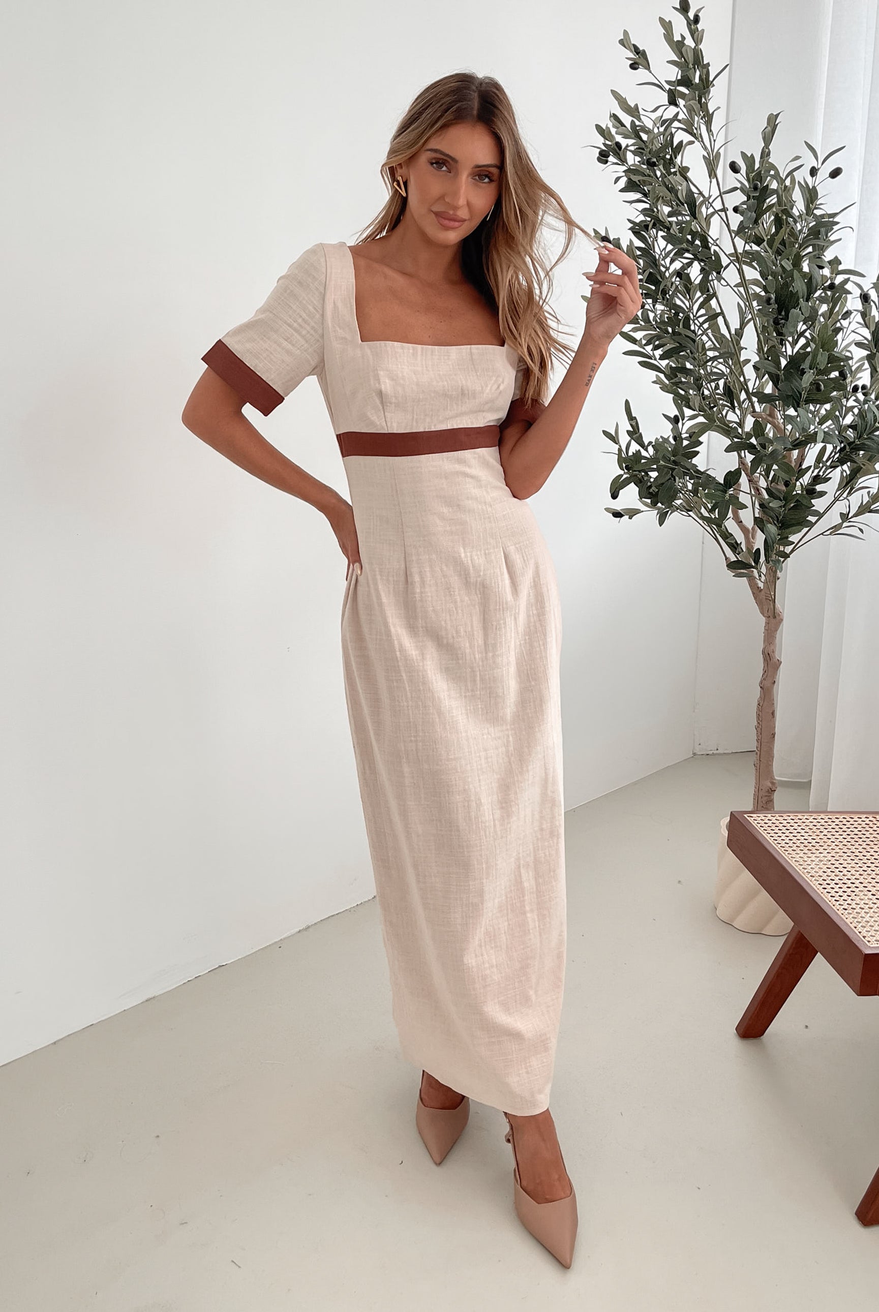 A woman with long, wavy blonde hair poses indoors in a minimalist room with white walls and a potted plant. She is wearing a light beige short-sleeve dress with brown trim details at the sleeves and waist, featuring a square neckline and a slim fit. She completes the look with pointed nude heels and a soft, confident expression.