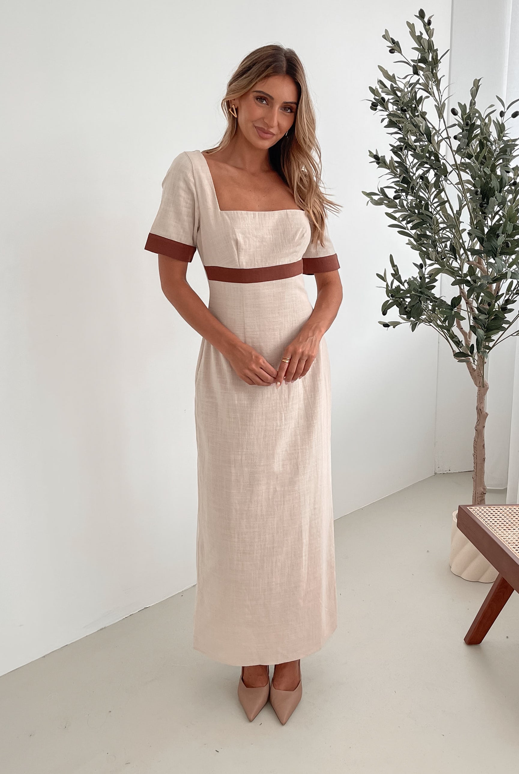 A woman with long, wavy blonde hair poses indoors in a minimalist room with white walls and a potted plant. She is wearing a light beige short-sleeve dress with brown trim details at the sleeves and waist, featuring a square neckline and a slim fit. She completes the look with pointed nude heels and a soft, confident expression.