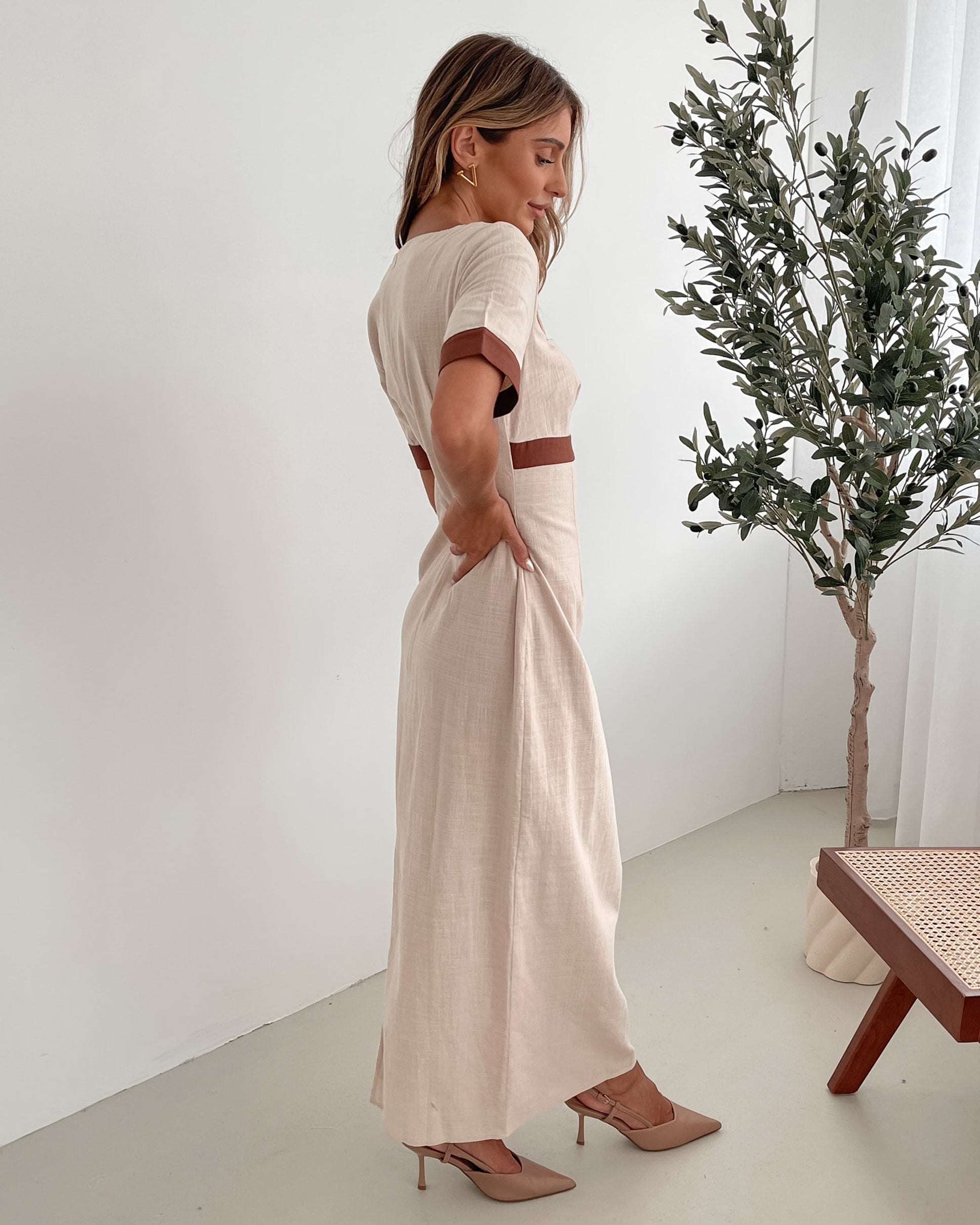A woman with long, wavy blonde hair poses indoors in a minimalist room with white walls and a potted plant. She is wearing a light beige short-sleeve dress with brown trim details at the sleeves and waist, featuring a square neckline and a slim fit. She completes the look with pointed nude heels and a soft, confident expression.