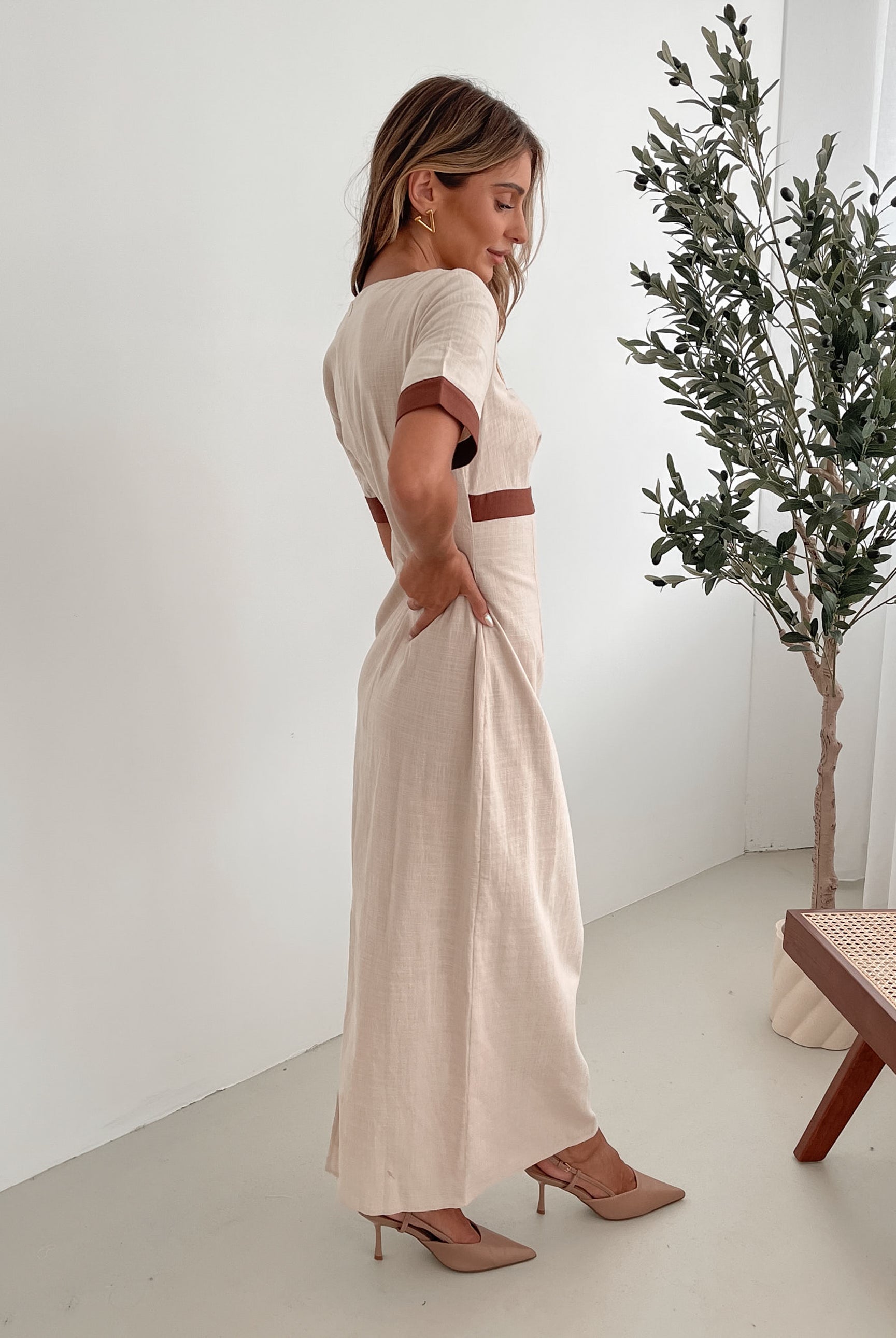 A woman with long, wavy blonde hair poses indoors in a minimalist room with white walls and a potted plant. She is wearing a light beige short-sleeve dress with brown trim details at the sleeves and waist, featuring a square neckline and a slim fit. She completes the look with pointed nude heels and a soft, confident expression.