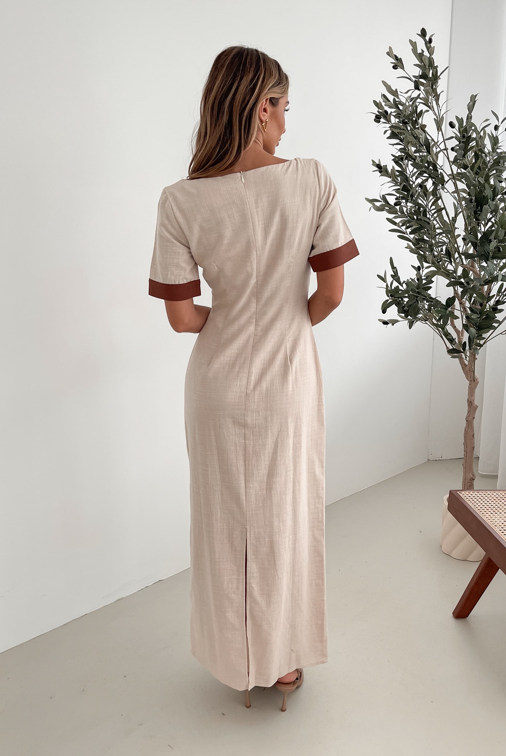 A woman with long, wavy blonde hair poses indoors in a minimalist room with white walls and a potted plant. She is wearing a light beige short-sleeve dress with brown trim details at the sleeves and waist, featuring a square neckline and a slim fit. She completes the look with pointed nude heels and a soft, confident expression.