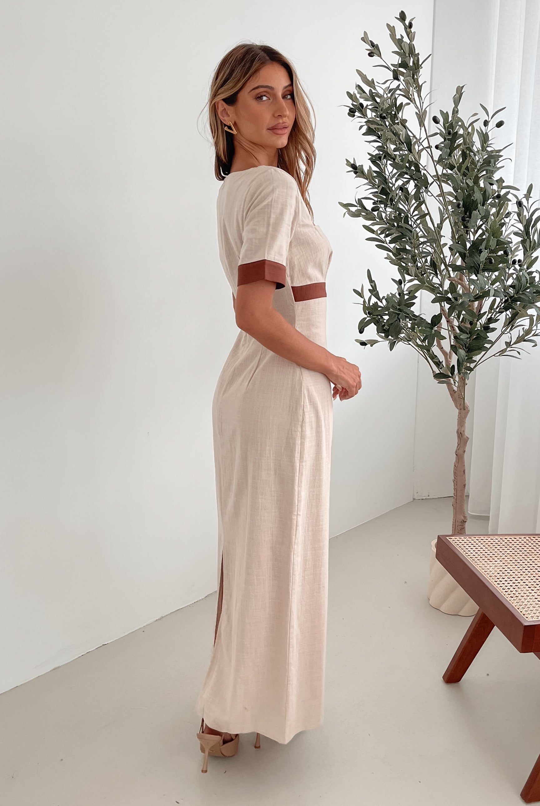 A woman with long, wavy blonde hair poses indoors in a minimalist room with white walls and a potted plant. She is wearing a light beige short-sleeve dress with brown trim details at the sleeves and waist, featuring a square neckline and a slim fit. She completes the look with pointed nude heels and a soft, confident expression.