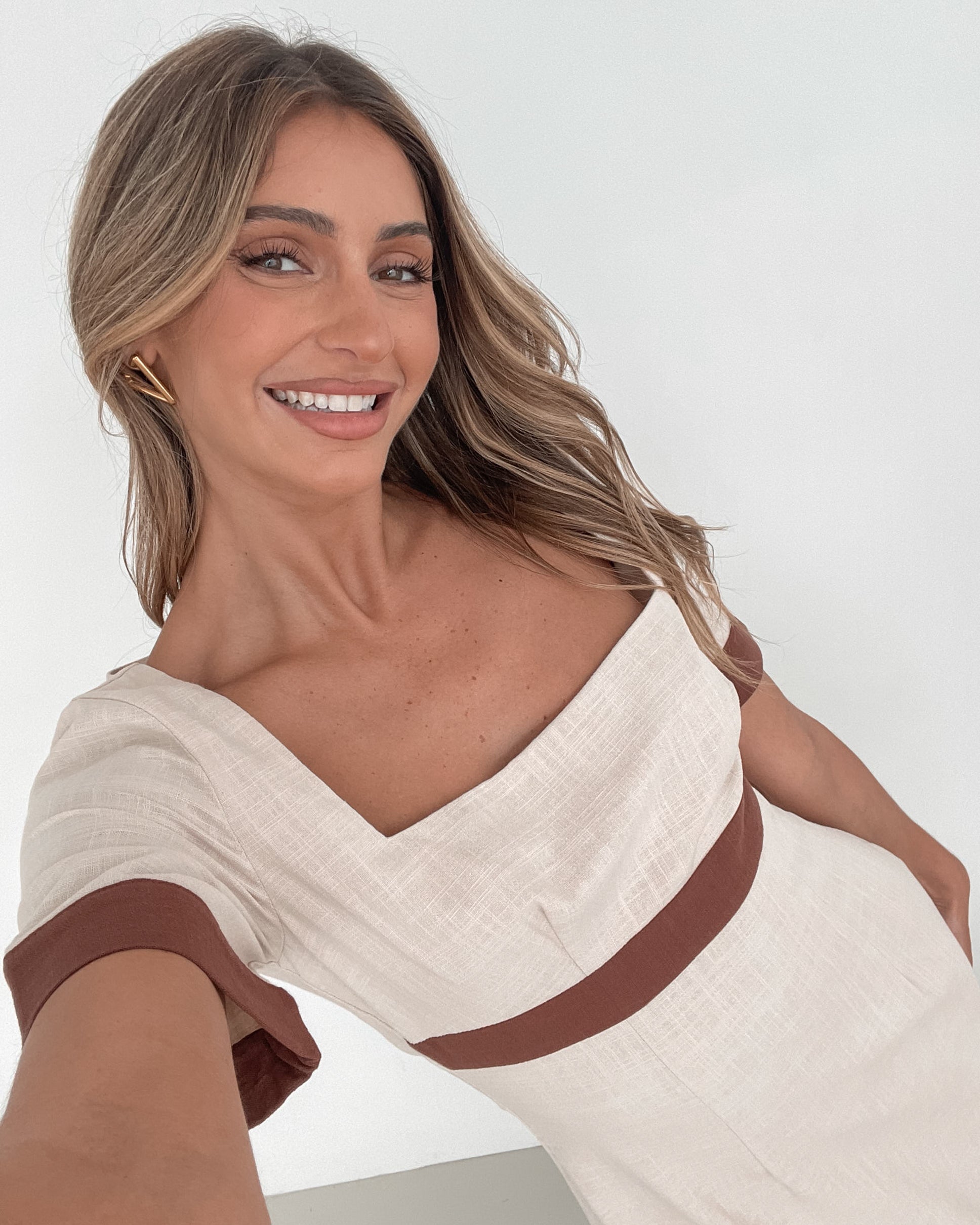A woman with long, wavy blonde hair poses indoors in a minimalist room with white walls and a potted plant. She is wearing a light beige short-sleeve dress with brown trim details at the sleeves and waist, featuring a square neckline and a slim fit. She completes the look with pointed nude heels and a soft, confident expression.