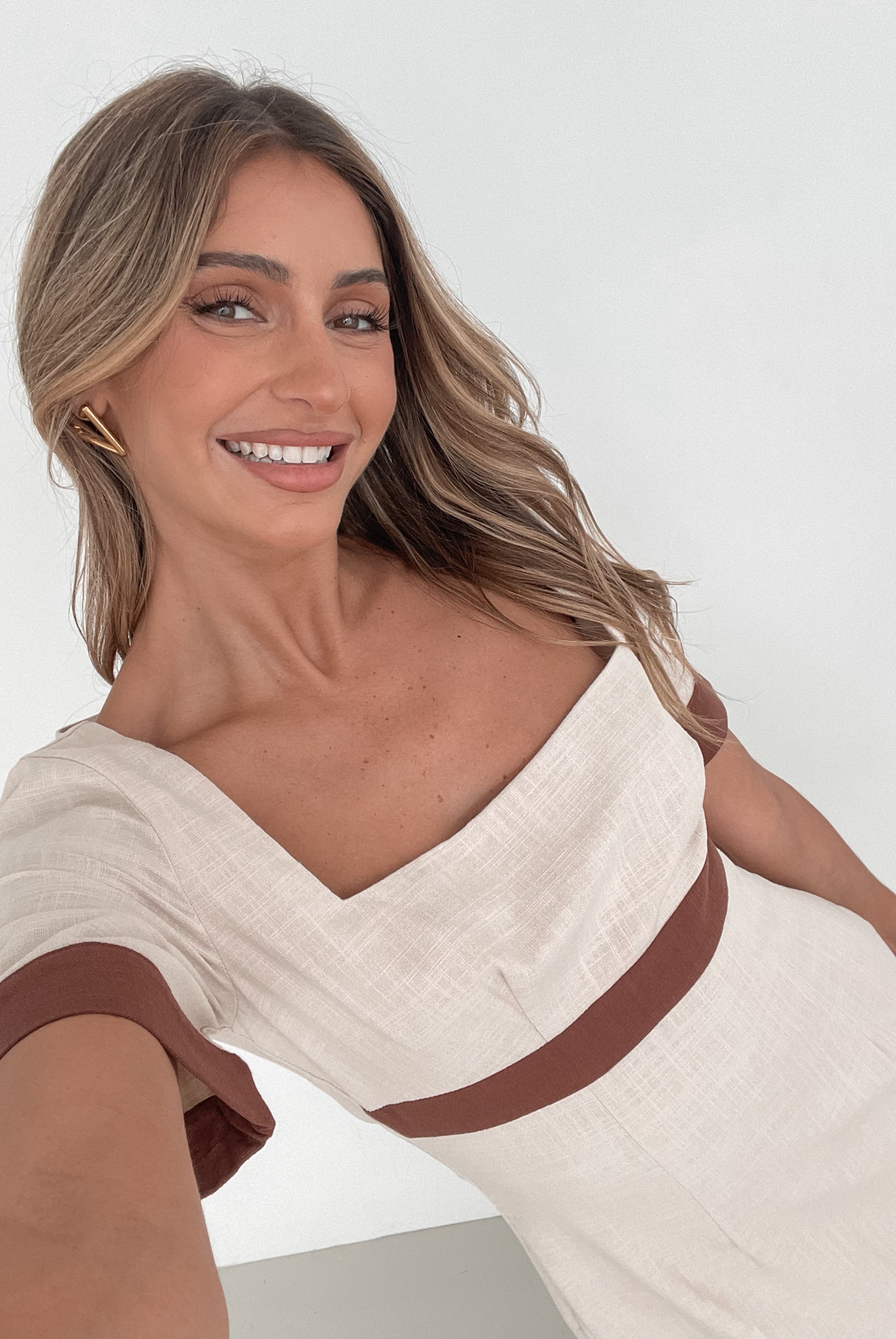 A woman with long, wavy blonde hair poses indoors in a minimalist room with white walls and a potted plant. She is wearing a light beige short-sleeve dress with brown trim details at the sleeves and waist, featuring a square neckline and a slim fit. She completes the look with pointed nude heels and a soft, confident expression.