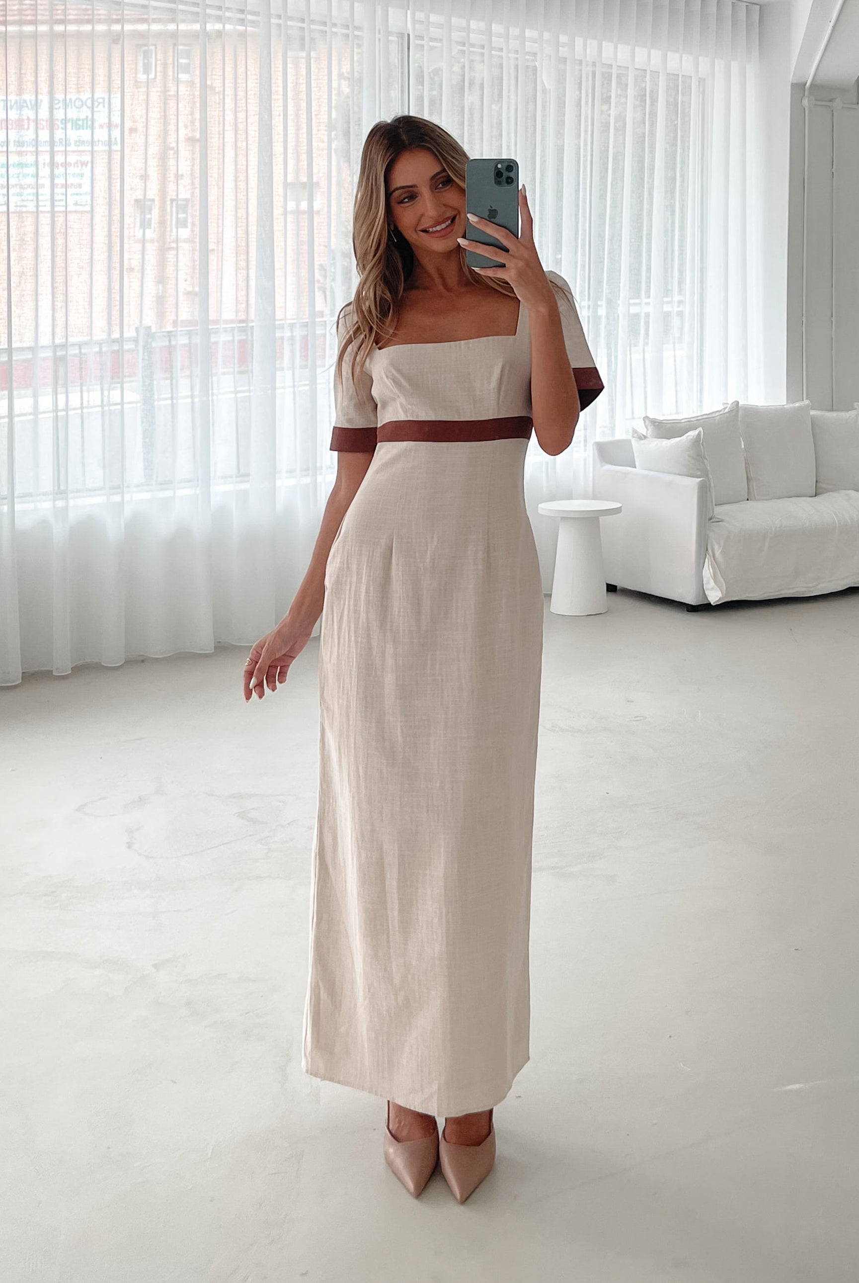 A woman with long, wavy blonde hair poses indoors in a minimalist room with white walls and a potted plant. She is wearing a light beige short-sleeve dress with brown trim details at the sleeves and waist, featuring a square neckline and a slim fit. She completes the look with pointed nude heels and a soft, confident expression.