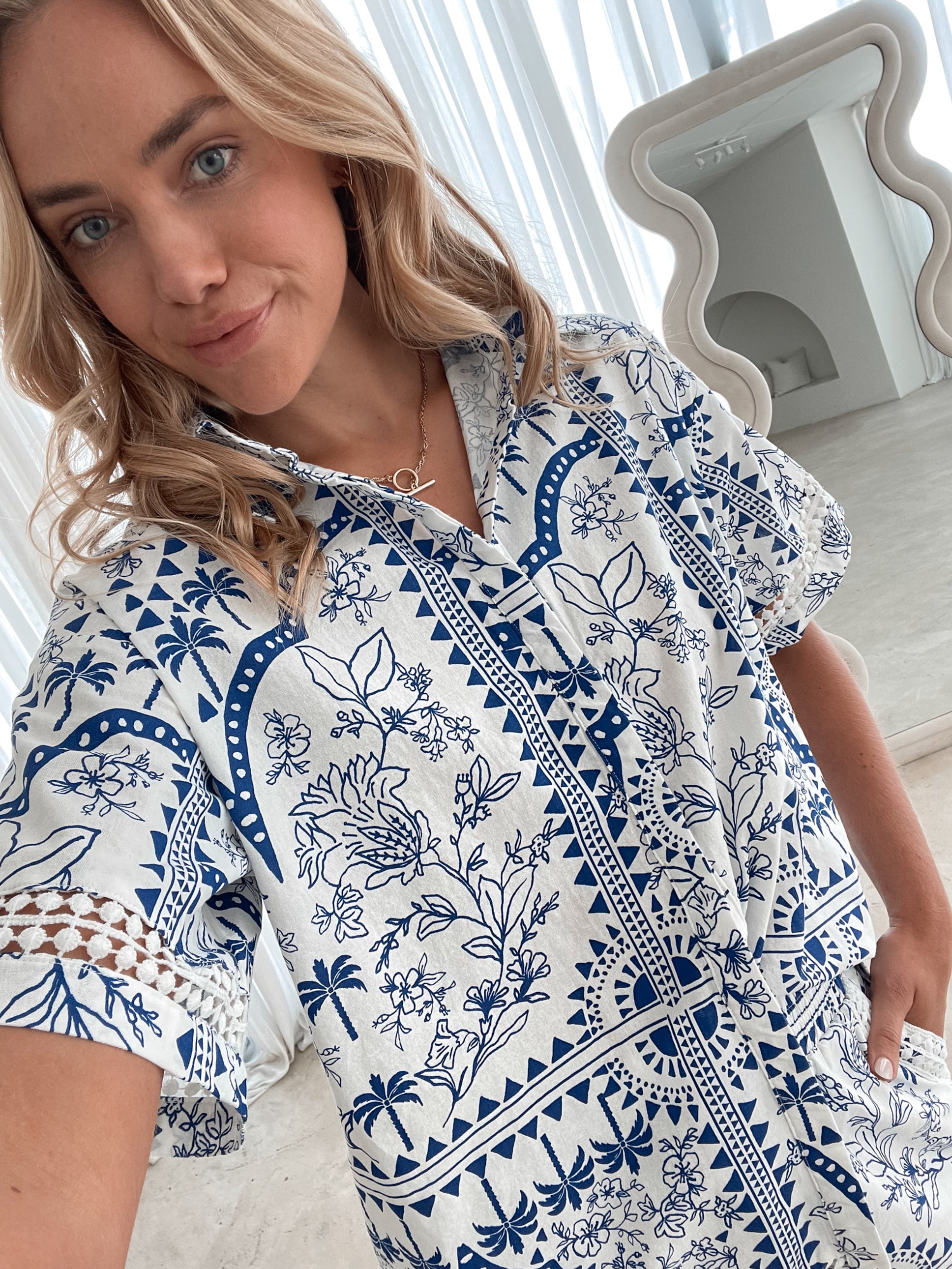 A woman with blonde, wavy hair stands in a bright, modern room with floor-to-ceiling sheer white curtains and neutral-toned decor. She is wearing a coordinated short-sleeve button-up shirt and matching shorts set featuring a blue and white floral and geometric print. She has a relaxed expression with one hand in her pocket, exuding a stylish and effortless vibe.