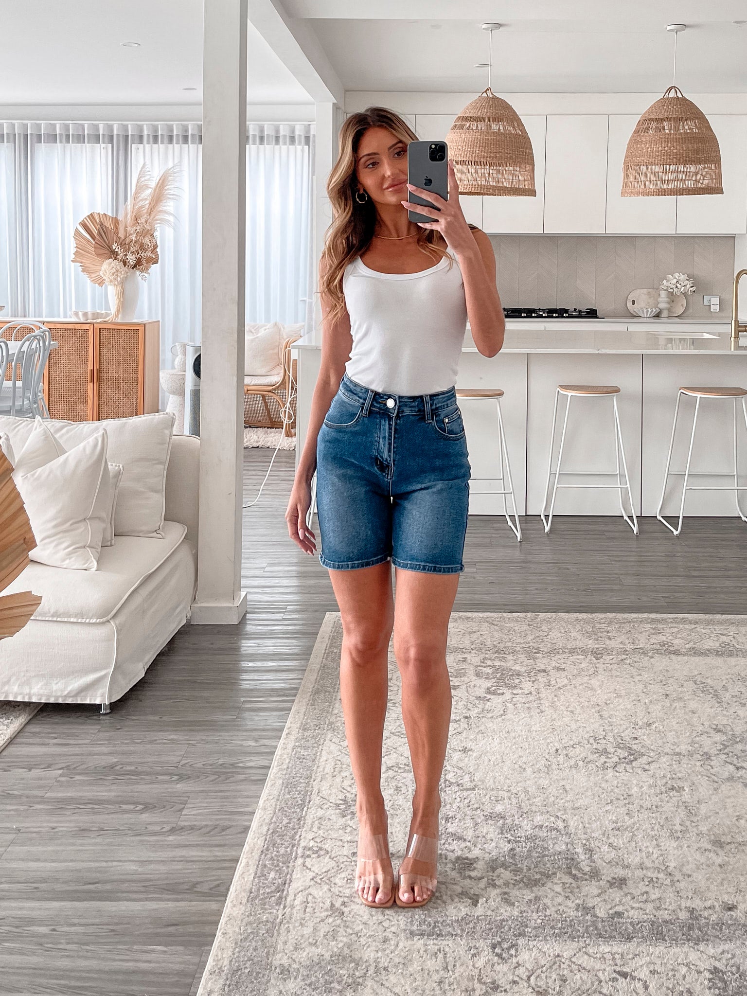 A smiling woman with long, wavy hair stands indoors in front of a white distressed wooden sideboard decorated with a large shell sculpture, a white vase holding dried palm leaves, and a round wall clock. She is wearing a white sleeveless tank top paired with high-waisted denim shorts and clear strap heels, with one hand resting in her pocket. The setting has a cozy, neutral-toned decor with a woven basket to the side and a patterned rug underfoot.