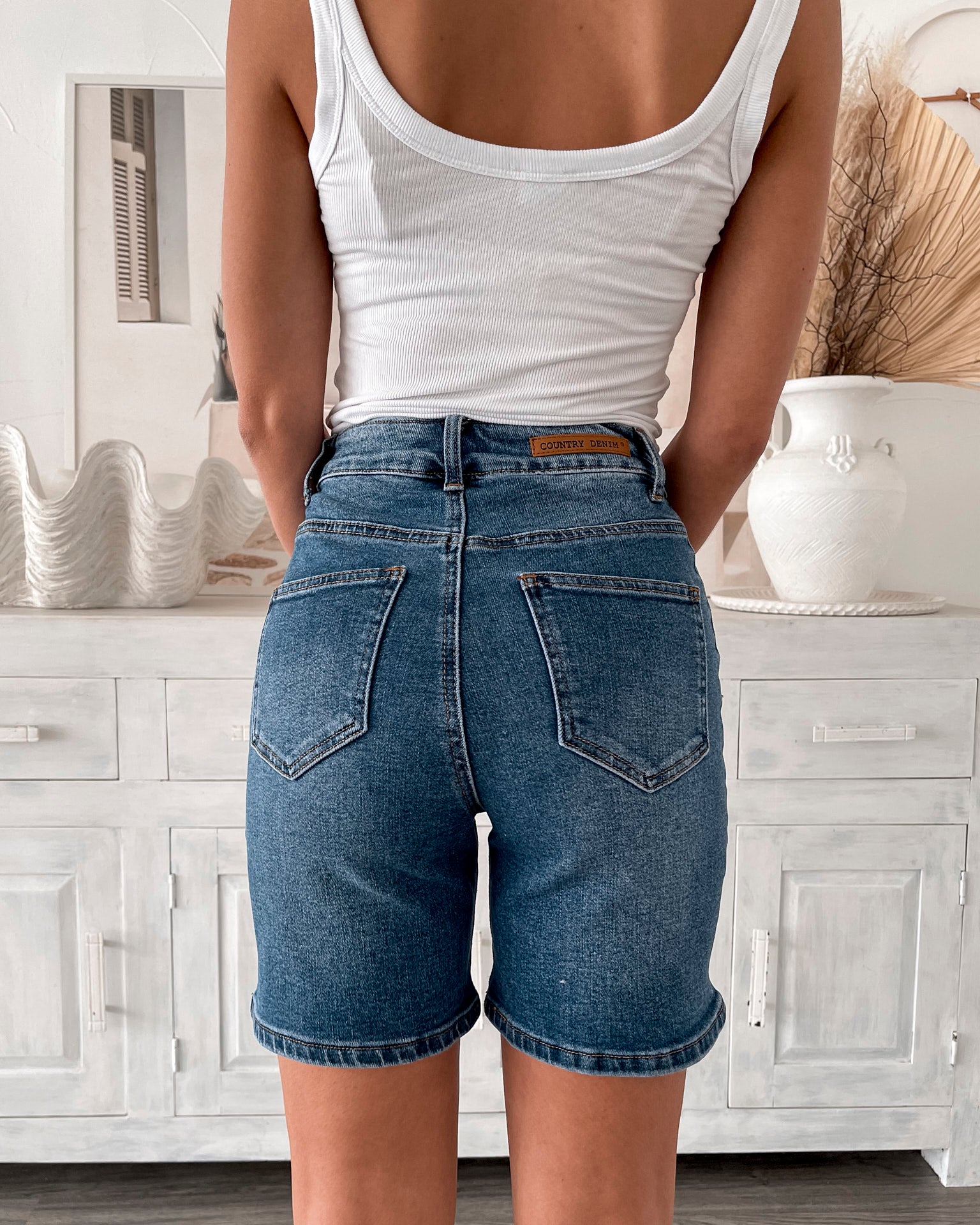 A smiling woman with long, wavy hair stands indoors in front of a white distressed wooden sideboard decorated with a large shell sculpture, a white vase holding dried palm leaves, and a round wall clock. She is wearing a white sleeveless tank top paired with high-waisted denim shorts and clear strap heels, with one hand resting in her pocket. The setting has a cozy, neutral-toned decor with a woven basket to the side and a patterned rug underfoot.