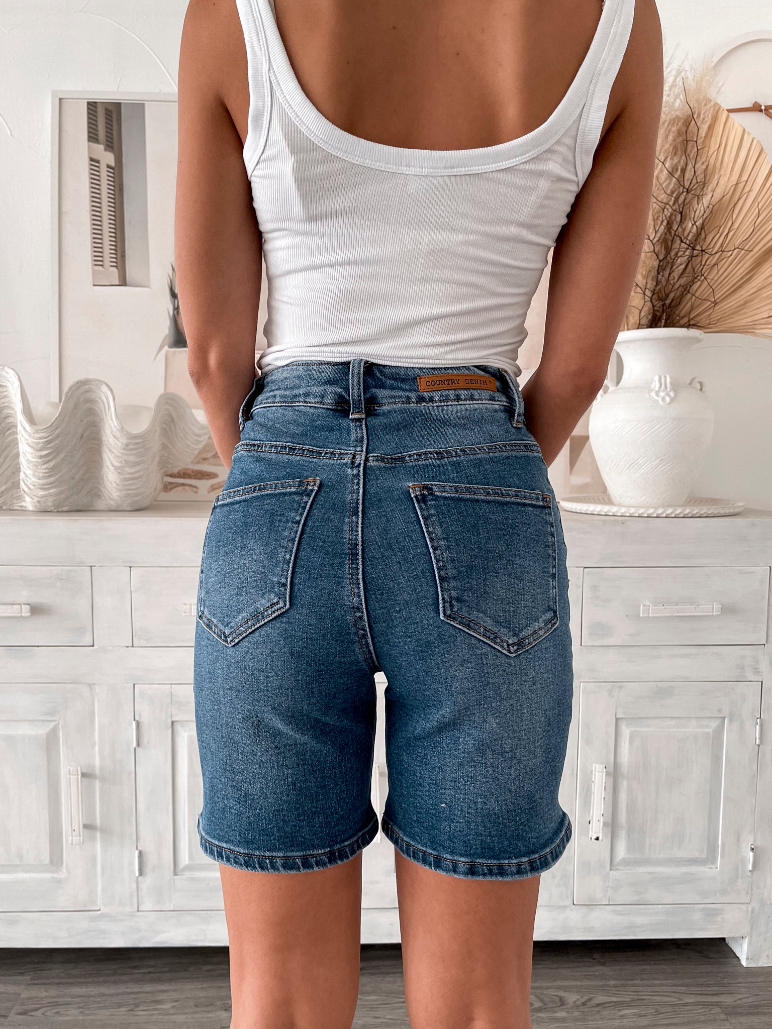 A smiling woman with long, wavy hair stands indoors in front of a white distressed wooden sideboard decorated with a large shell sculpture, a white vase holding dried palm leaves, and a round wall clock. She is wearing a white sleeveless tank top paired with high-waisted denim shorts and clear strap heels, with one hand resting in her pocket. The setting has a cozy, neutral-toned decor with a woven basket to the side and a patterned rug underfoot.