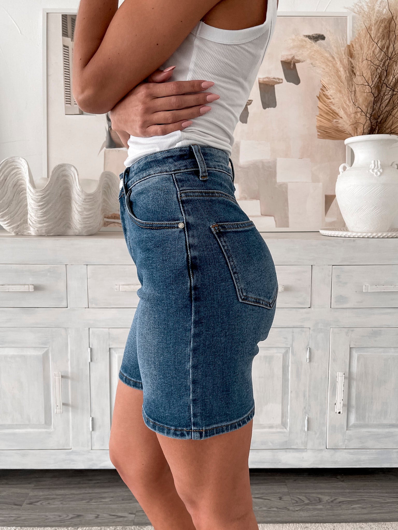 A smiling woman with long, wavy hair stands indoors in front of a white distressed wooden sideboard decorated with a large shell sculpture, a white vase holding dried palm leaves, and a round wall clock. She is wearing a white sleeveless tank top paired with high-waisted denim shorts and clear strap heels, with one hand resting in her pocket. The setting has a cozy, neutral-toned decor with a woven basket to the side and a patterned rug underfoot.