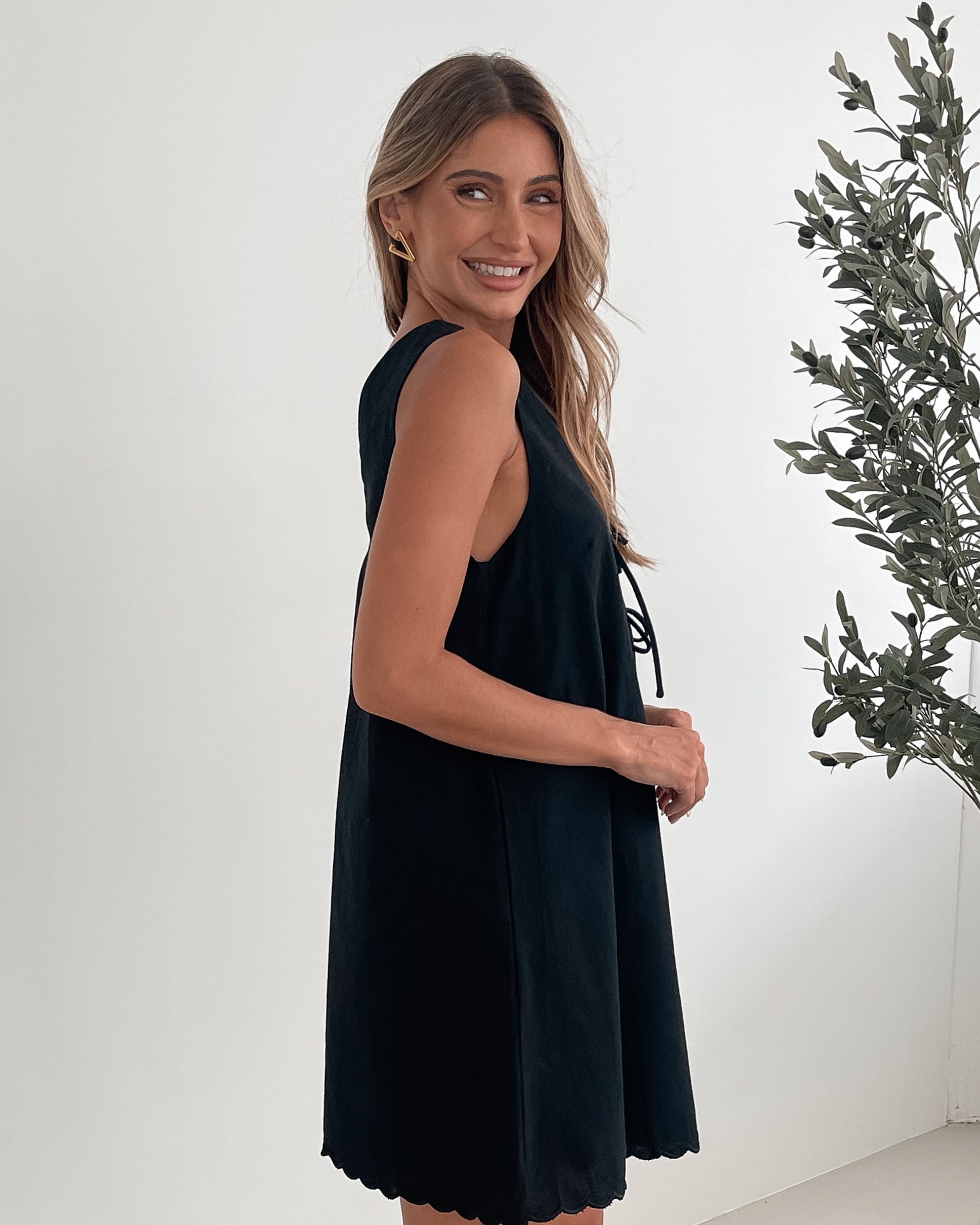 A woman with long, wavy blonde hair stands indoors against a plain white wall with a green leafy plant to her side. She is wearing a sleeveless black dress with a V-neck and two front tie details, featuring a scalloped hem. Her left hand rests on her hip while her right hand gently touches the side of the dress. She has a calm expression and wears subtle makeup and small hoop earrings.