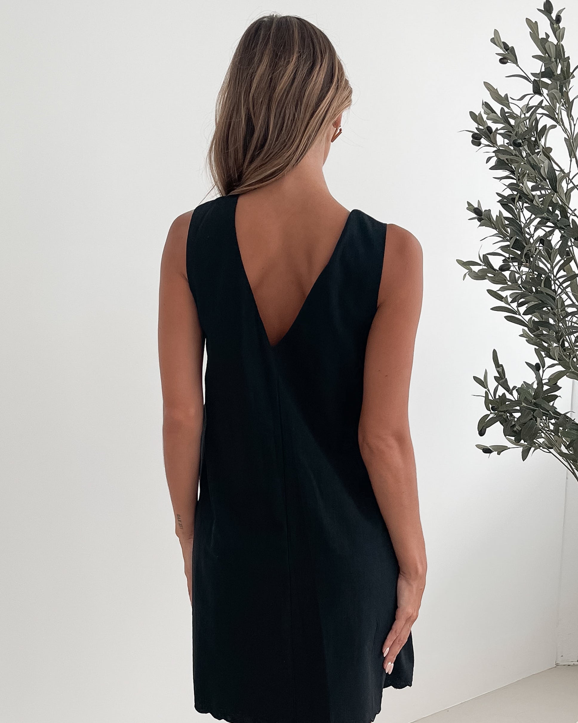 A woman with long, wavy blonde hair stands indoors against a plain white wall with a green leafy plant to her side. She is wearing a sleeveless black dress with a V-neck and two front tie details, featuring a scalloped hem. Her left hand rests on her hip while her right hand gently touches the side of the dress. She has a calm expression and wears subtle makeup and small hoop earrings.