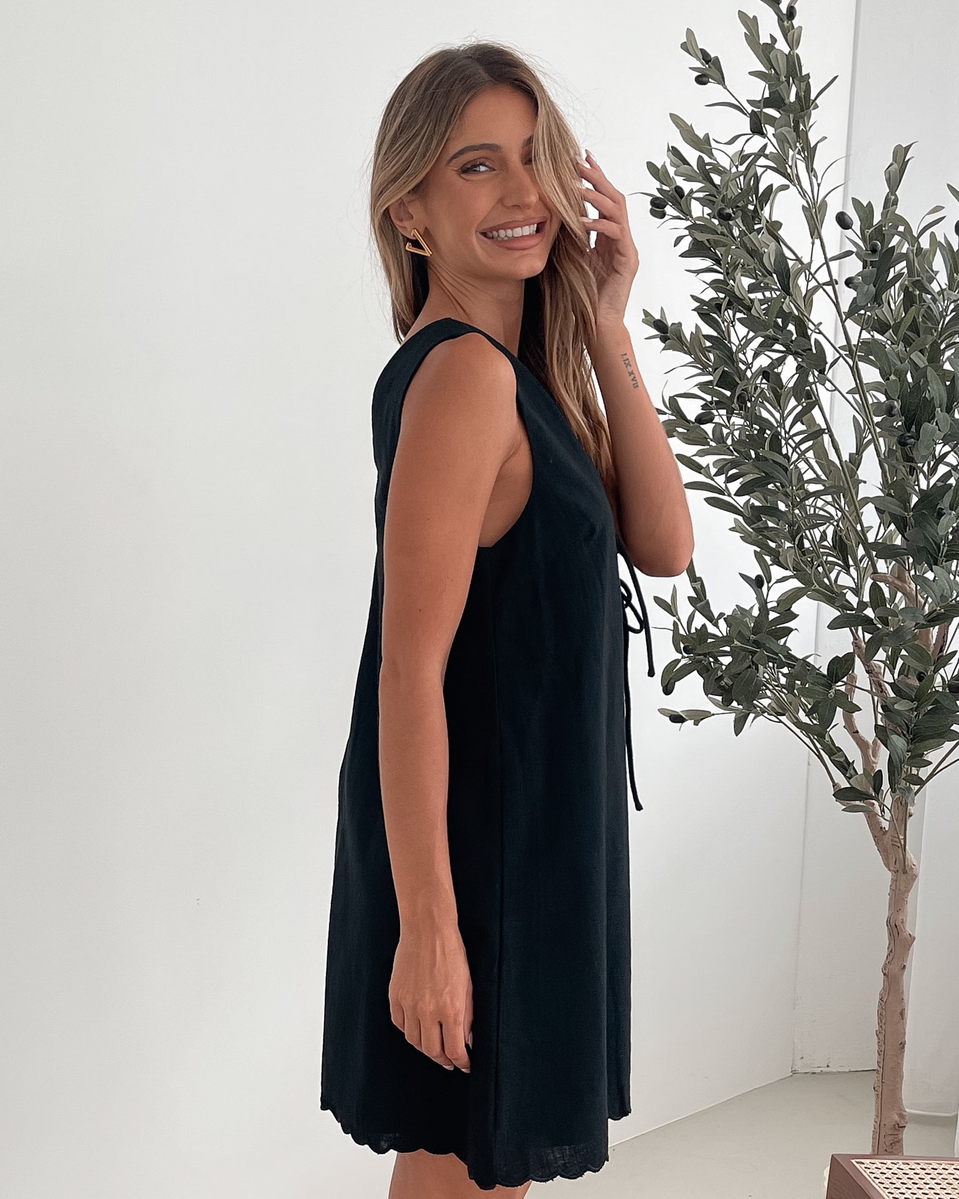 A woman with long, wavy blonde hair stands indoors against a plain white wall with a green leafy plant to her side. She is wearing a sleeveless black dress with a V-neck and two front tie details, featuring a scalloped hem. Her left hand rests on her hip while her right hand gently touches the side of the dress. She has a calm expression and wears subtle makeup and small hoop earrings.