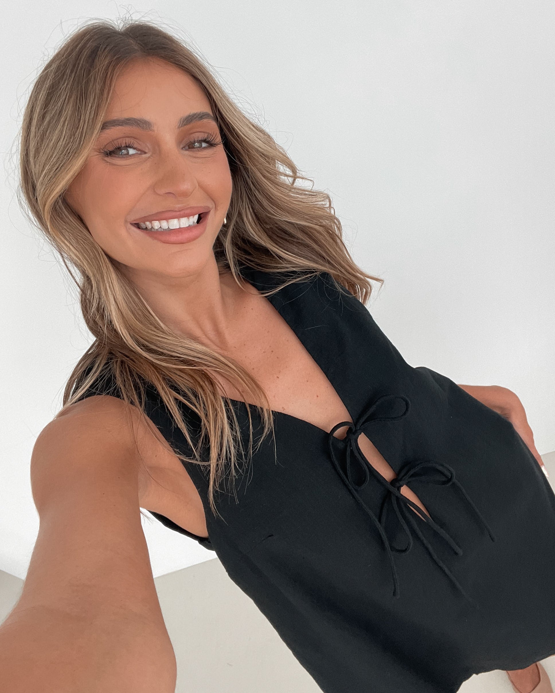 A woman with long, wavy blonde hair stands indoors against a plain white wall with a green leafy plant to her side. She is wearing a sleeveless black dress with a V-neck and two front tie details, featuring a scalloped hem. Her left hand rests on her hip while her right hand gently touches the side of the dress. She has a calm expression and wears subtle makeup and small hoop earrings.