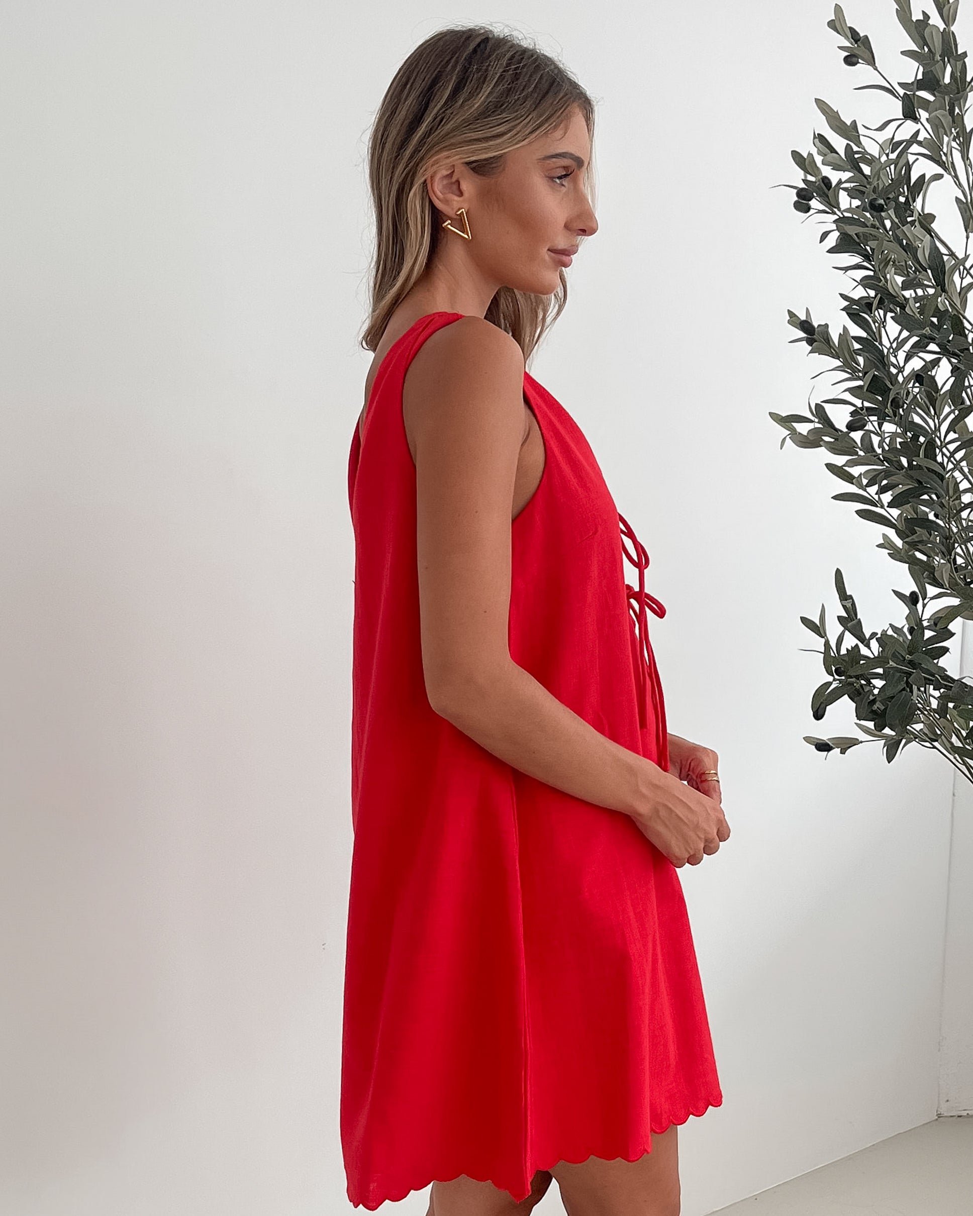 A woman with long, wavy blonde hair stands indoors in front of a plain white wall, with a green leafy plant beside her. She is wearing a sleeveless red dress with a V-neck and two front tie details, similar in style to the previous dress, featuring a scalloped hem. Her hands are gently clasped together in front of her, and she has a subtle smile on her face. She accessorizes with gold hoop earrings and a delicate ring, and her makeup is natural and polished.