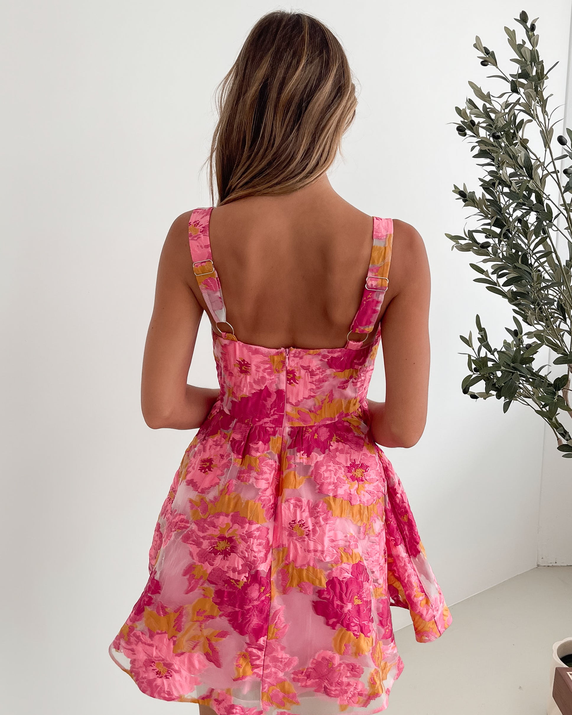 A young woman with long, wavy blonde hair stands indoors against a simple white wall, with a tall leafy plant nearby. She is wearing a sleeveless, strapless summer dress featuring a vibrant floral pattern in shades of pink, magenta, and yellow. The dress has a fitted bodice with a square neckline and a flared skirt that adds a playful and feminine touch. She has a soft smile and her right hand is gently touching her hair, exuding a cheerful and relaxed vibe.
