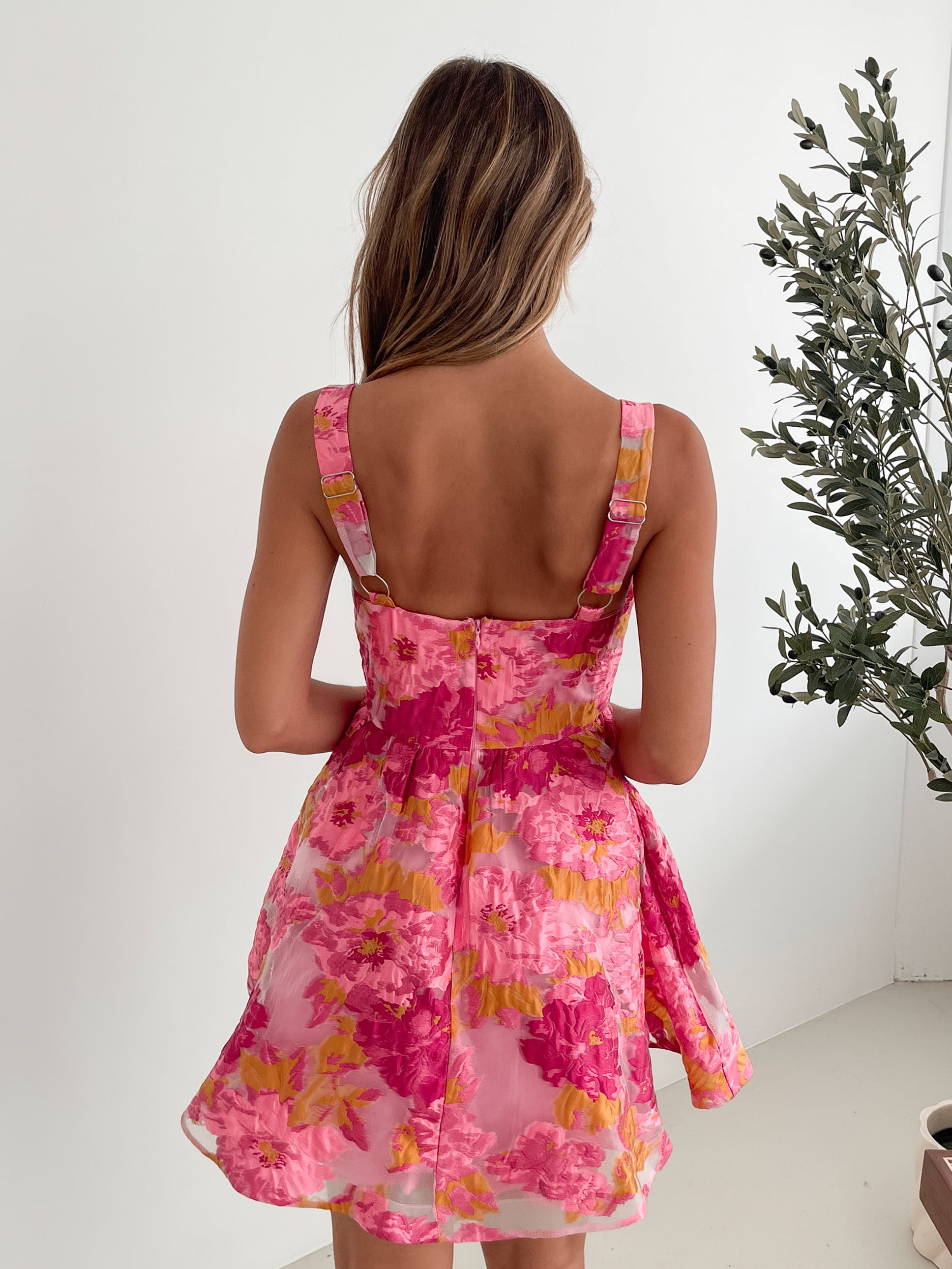 A young woman with long, wavy blonde hair stands indoors against a simple white wall, with a tall leafy plant nearby. She is wearing a sleeveless, strapless summer dress featuring a vibrant floral pattern in shades of pink, magenta, and yellow. The dress has a fitted bodice with a square neckline and a flared skirt that adds a playful and feminine touch. She has a soft smile and her right hand is gently touching her hair, exuding a cheerful and relaxed vibe.