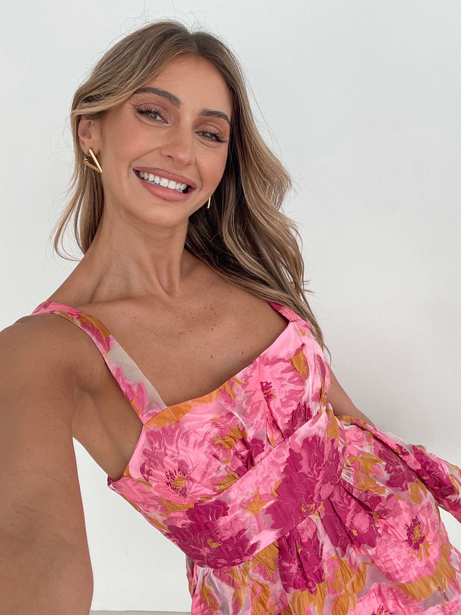 A young woman with long, wavy blonde hair stands indoors against a simple white wall, with a tall leafy plant nearby. She is wearing a sleeveless, strapless summer dress featuring a vibrant floral pattern in shades of pink, magenta, and yellow. The dress has a fitted bodice with a square neckline and a flared skirt that adds a playful and feminine touch. She has a soft smile and her right hand is gently touching her hair, exuding a cheerful and relaxed vibe.