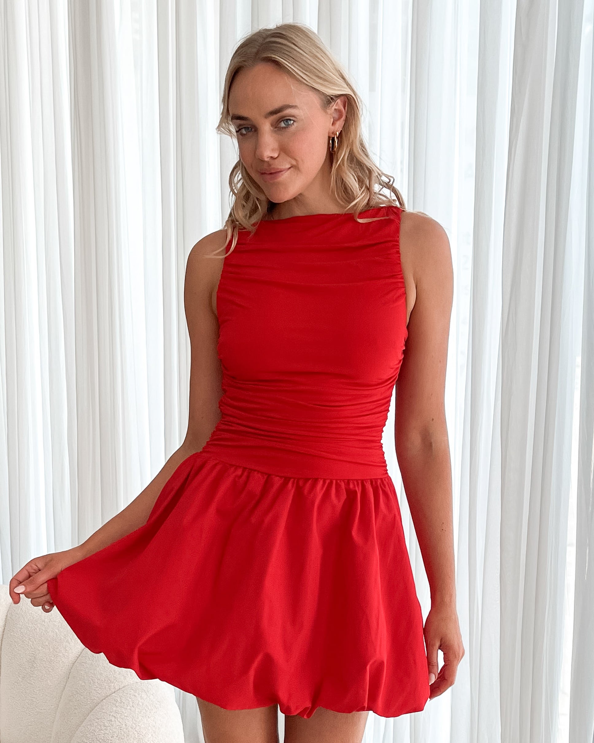 A young woman with blonde hair styled in loose waves stands indoors in front of sheer white curtains. She is wearing a vibrant red sleeveless dress with a fitted, ruched bodice and a playful, bubble-hem skirt. The dress has a high neckline and accentuates her waist, creating a flattering silhouette. She holds the skirt slightly out with one hand, smiling softly with a confident and relaxed expression. The setting and her pose give off an elegant yet fun and youthful vibe.