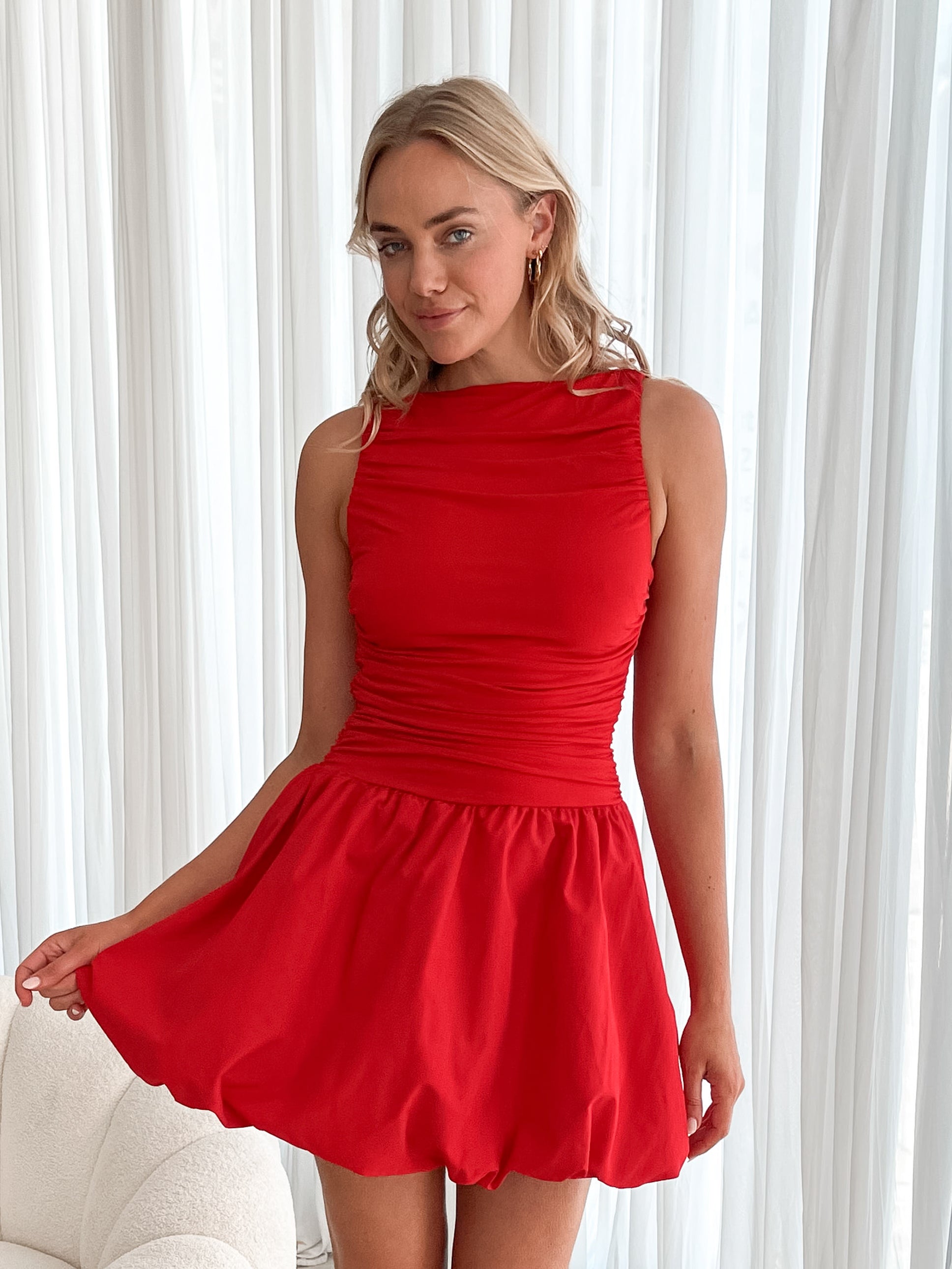 A young woman with blonde hair styled in loose waves stands indoors in front of sheer white curtains. She is wearing a vibrant red sleeveless dress with a fitted, ruched bodice and a playful, bubble-hem skirt. The dress has a high neckline and accentuates her waist, creating a flattering silhouette. She holds the skirt slightly out with one hand, smiling softly with a confident and relaxed expression. The setting and her pose give off an elegant yet fun and youthful vibe.