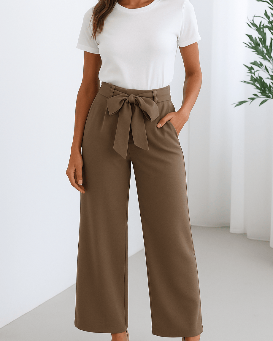 A woman wearing a white short-sleeve t-shirt tucked into high-waisted, wide-leg brown trousers with a waist tie belt, standing in a minimalistic room with light walls and a green plant in the background.