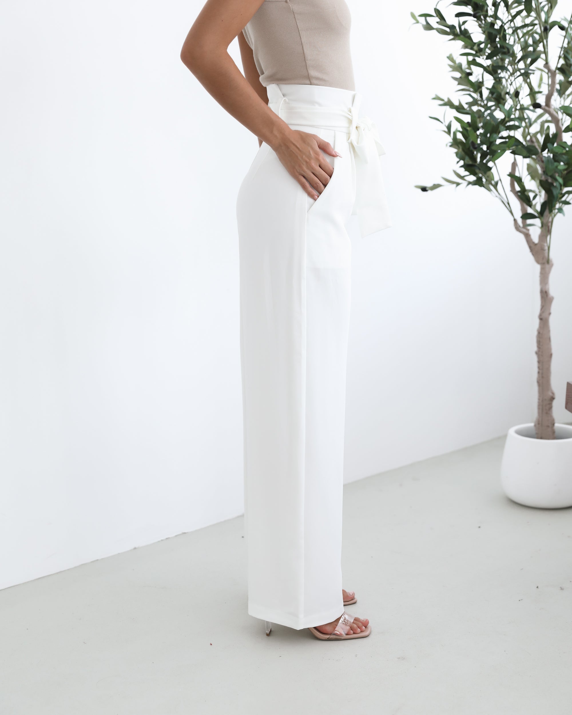 A woman wearing high-waisted, wide-leg white trousers with a paper bag waist and a fabric tie belt, paired with a sleeveless beige ribbed top and nude heels, standing in a minimalistic room with a potted tree and a wooden bench.