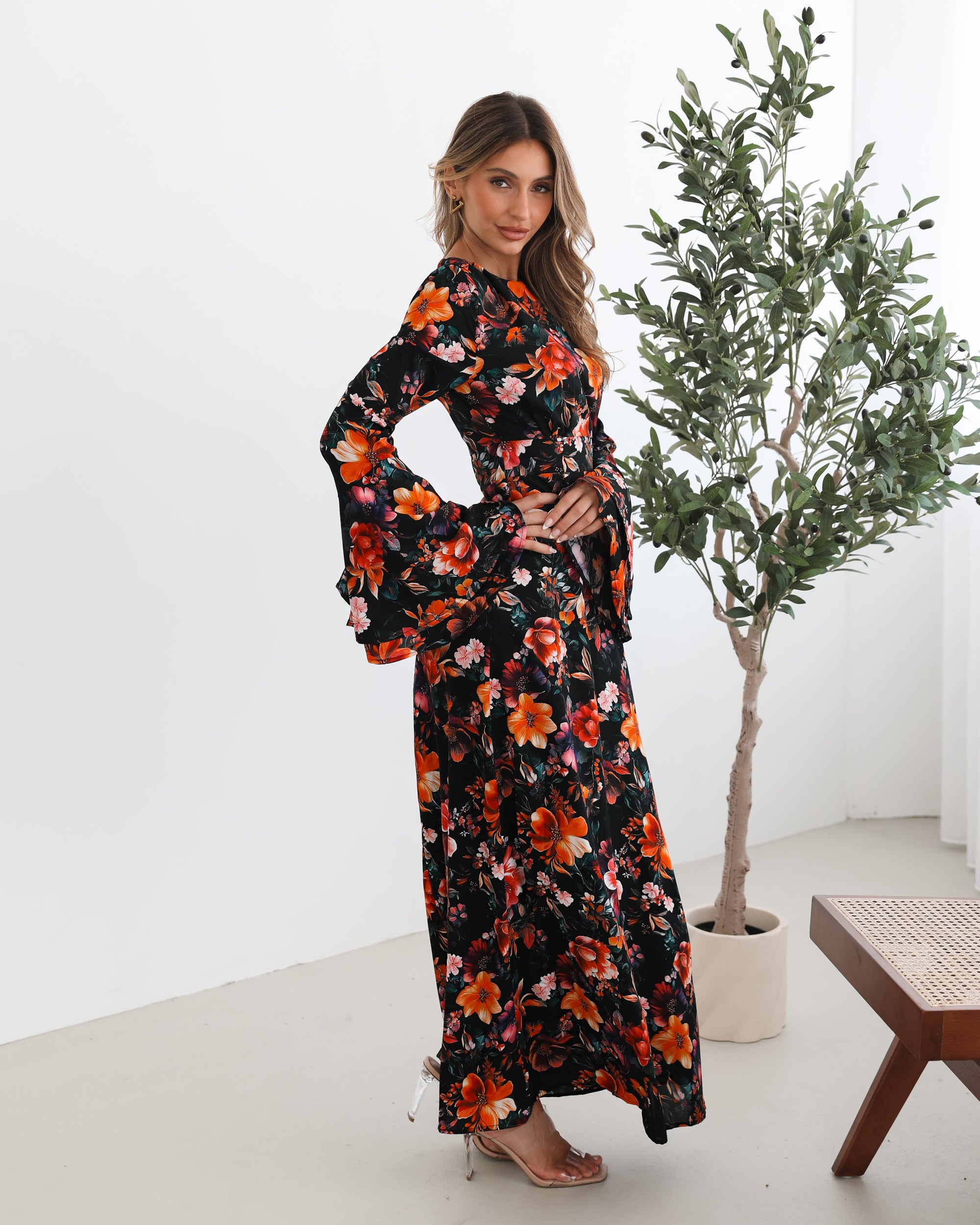 A woman wearing a long, black floral maxi dress with vibrant orange, red, and pink flowers. The dress features long bell sleeves and a fitted waist. She is leaning against a white wall in a minimalistic room with a potted plant nearby.