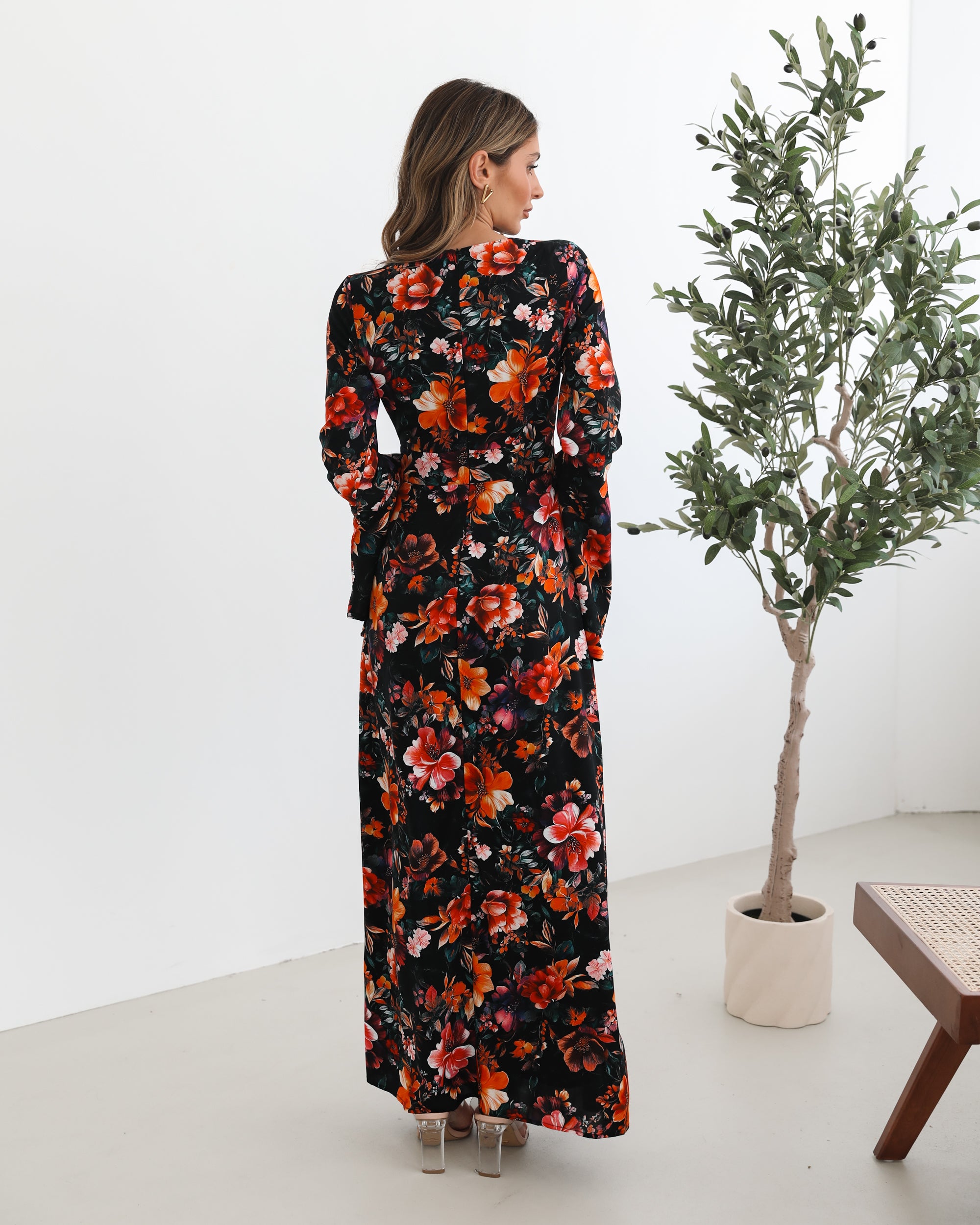 A woman wearing a long, black floral maxi dress with vibrant orange, red, and pink flowers. The dress features long bell sleeves and a fitted waist. She is leaning against a white wall in a minimalistic room with a potted plant nearby.
