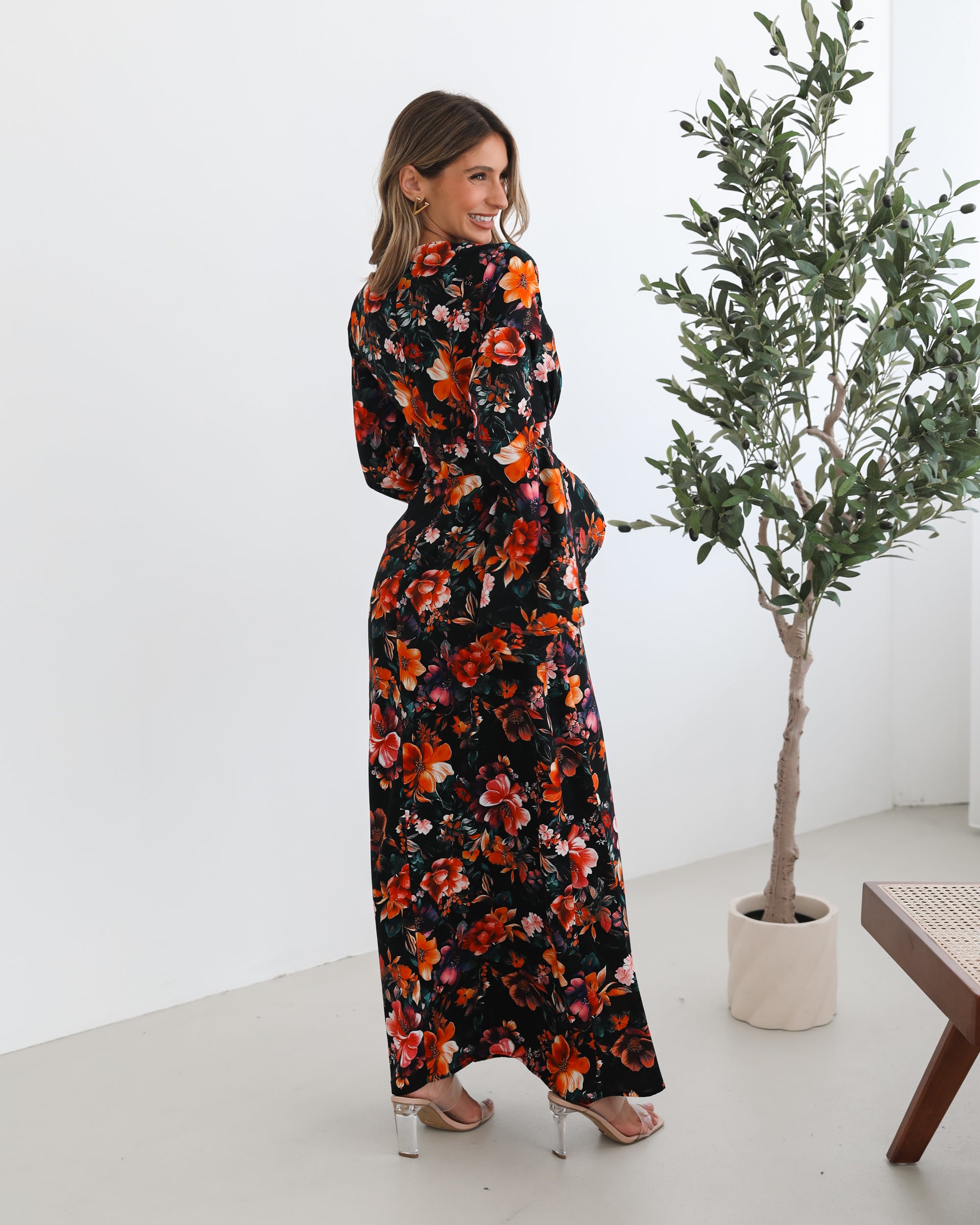 A woman wearing a long, black floral maxi dress with vibrant orange, red, and pink flowers. The dress features long bell sleeves and a fitted waist. She is leaning against a white wall in a minimalistic room with a potted plant nearby.