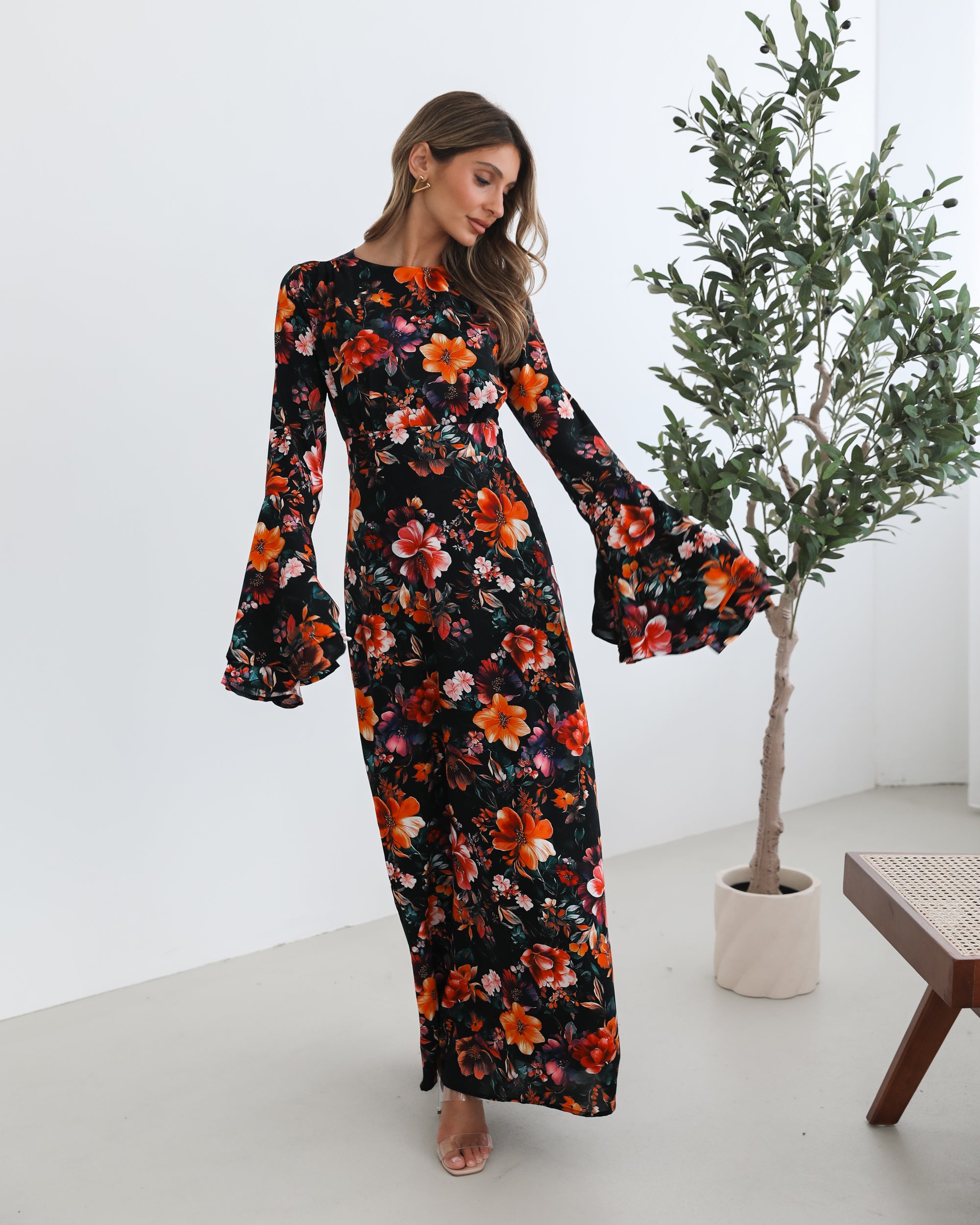 A woman wearing a long, black floral maxi dress with vibrant orange, red, and pink flowers. The dress features long bell sleeves and a fitted waist. She is leaning against a white wall in a minimalistic room with a potted plant nearby.