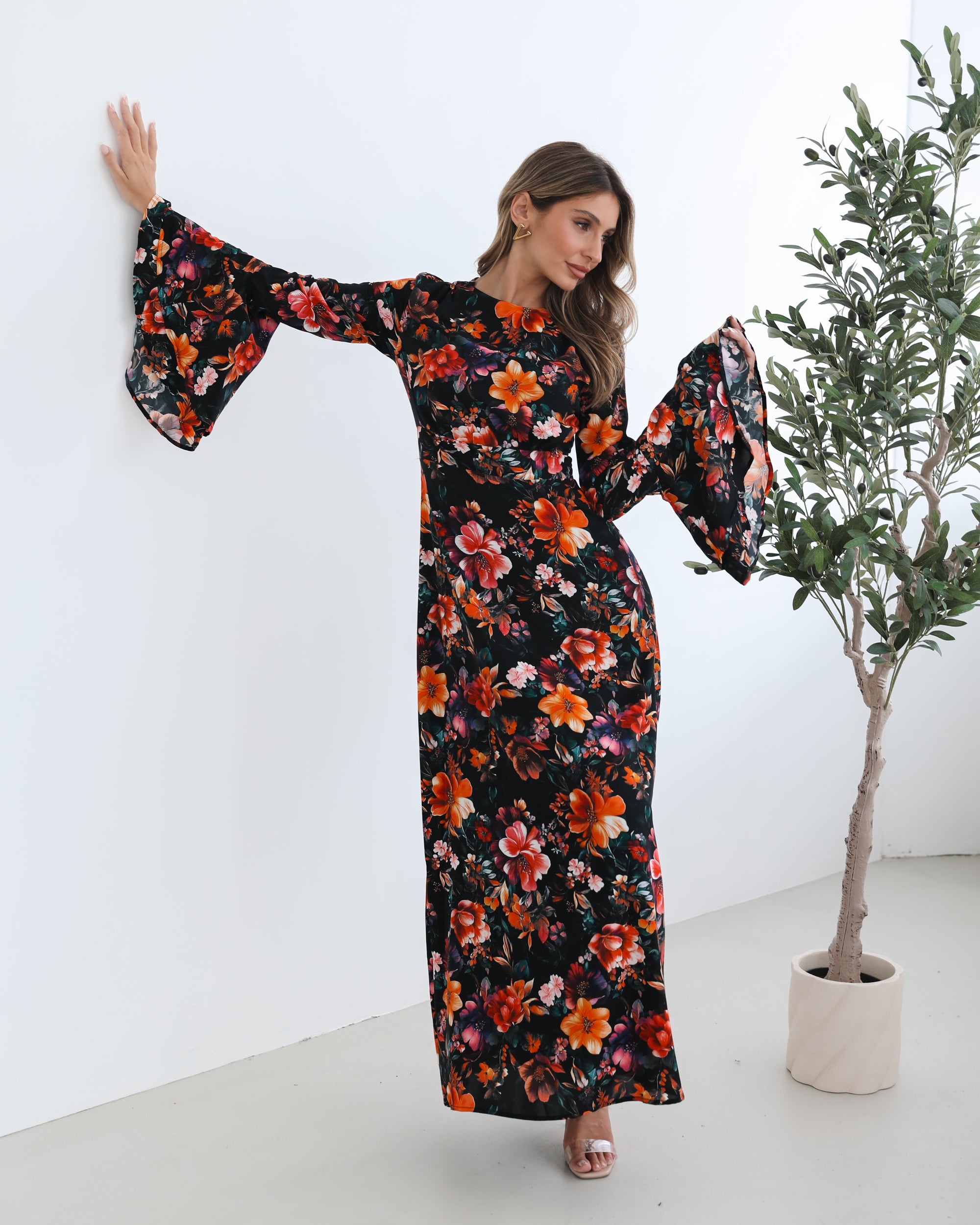 A woman wearing a long, black floral maxi dress with vibrant orange, red, and pink flowers. The dress features long bell sleeves and a fitted waist. She is leaning against a white wall in a minimalistic room with a potted plant nearby.