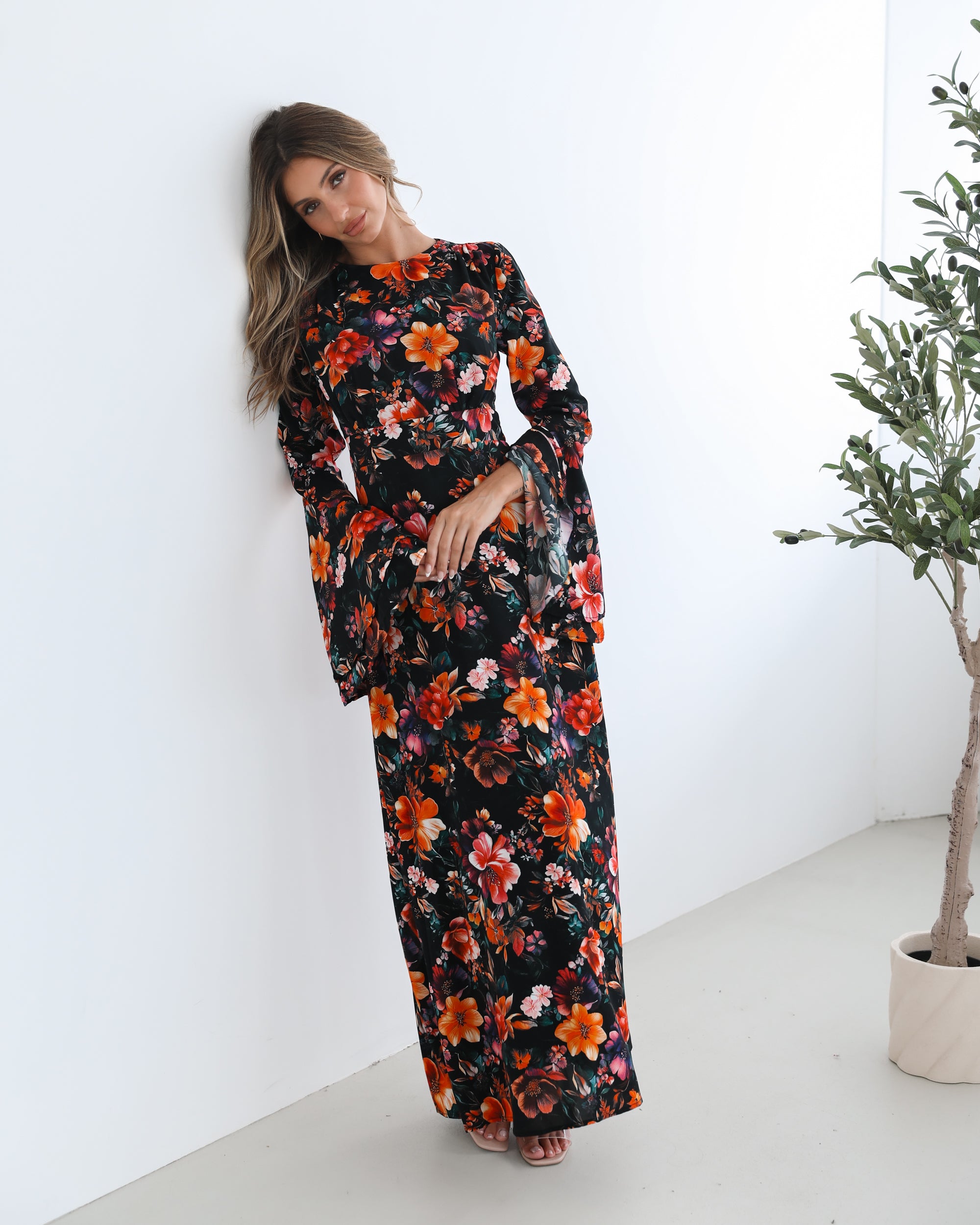 A woman wearing a long, black floral maxi dress with vibrant orange, red, and pink flowers. The dress features long bell sleeves and a fitted waist. She is leaning against a white wall in a minimalistic room with a potted plant nearby.