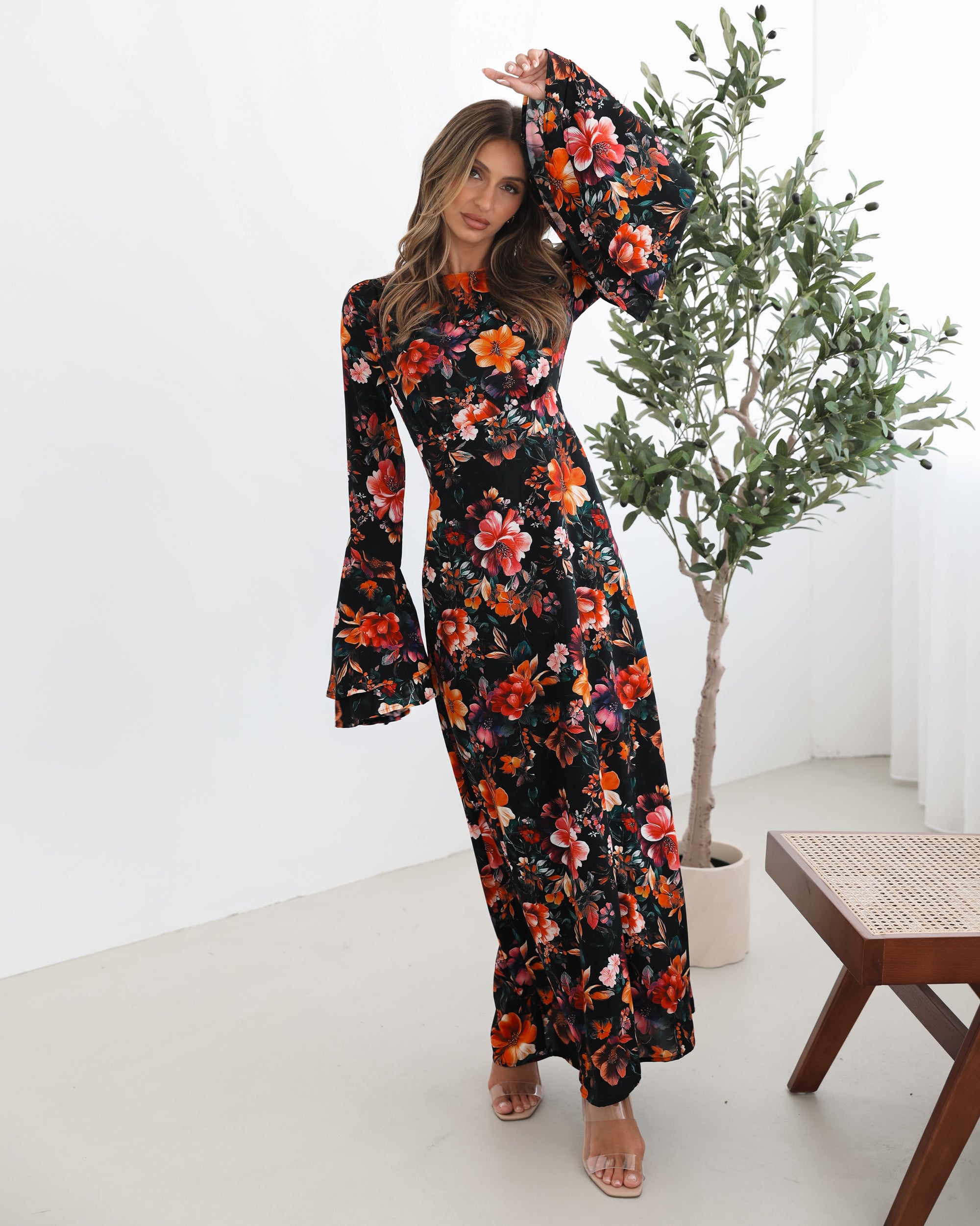 A woman wearing a long, black floral maxi dress with vibrant orange, red, and pink flowers. The dress features long bell sleeves and a fitted waist. She is leaning against a white wall in a minimalistic room with a potted plant nearby.