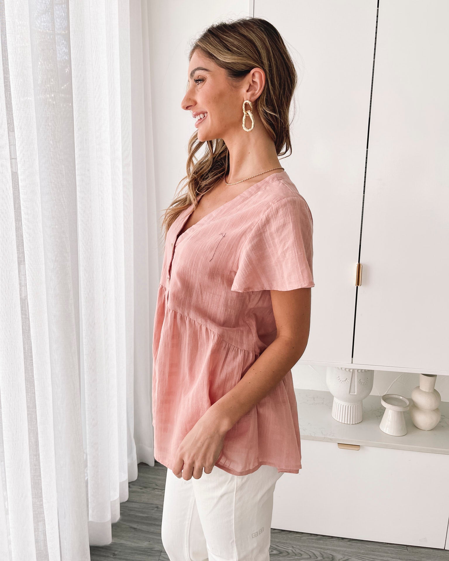 Smiling young woman with long blonde hair wearing a light pink button-up blouse and white shorts, standing indoors next to a white desk and wicker chair, with decorative art pieces in the background.