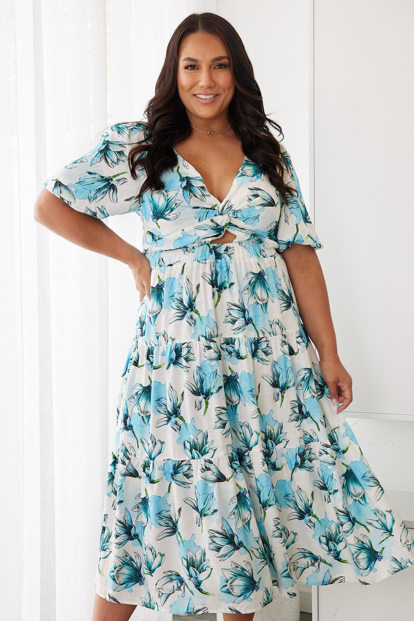 Woman wearing a white and blue floral midi dress with a front cut-out and ruched detailing, featuring elbow-length sleeves and a flowy skirt, standing indoors and smiling.