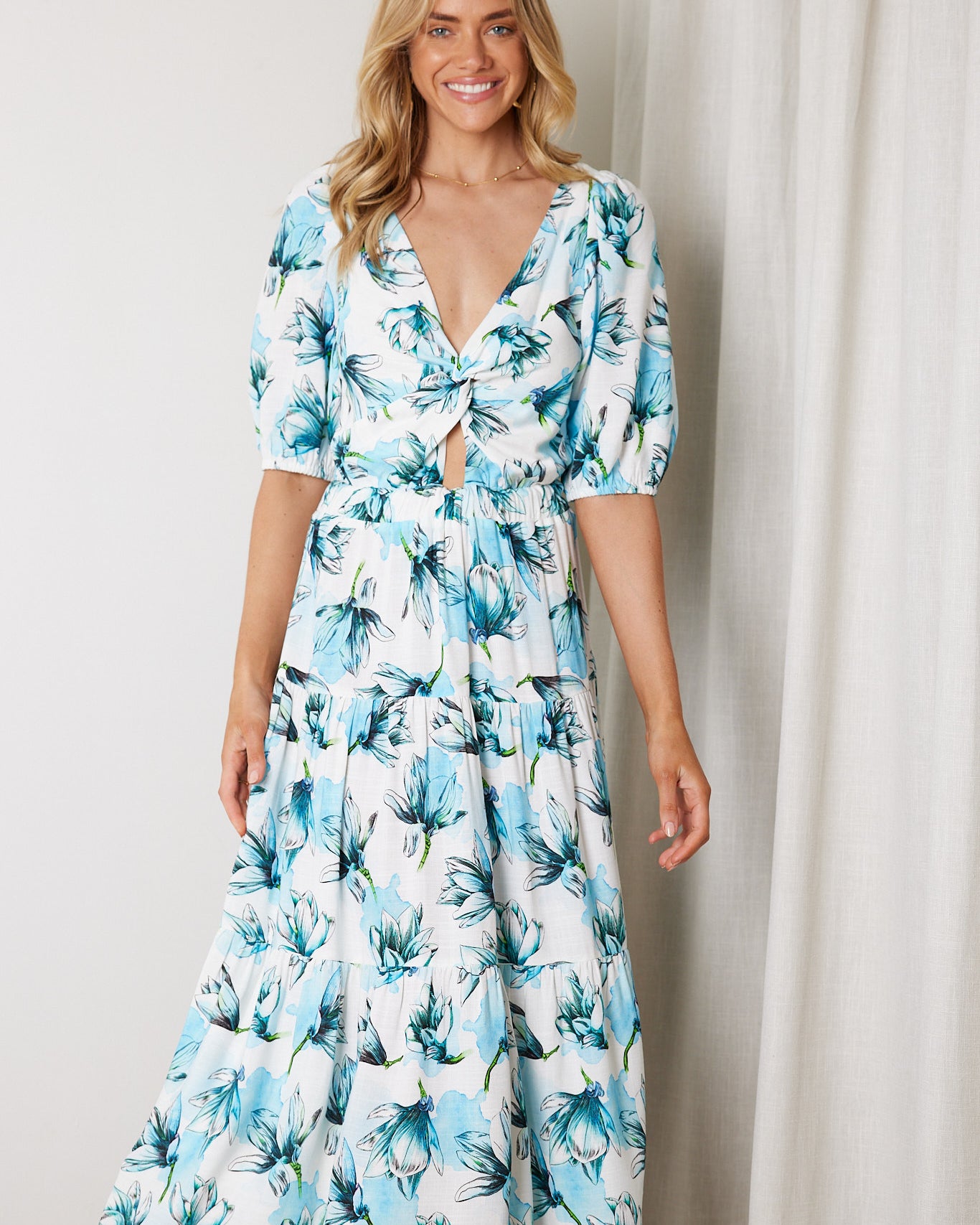 Woman wearing a white and blue floral midi dress with a front cut-out and ruched detailing, featuring elbow-length sleeves and a flowy skirt, standing indoors and smiling.