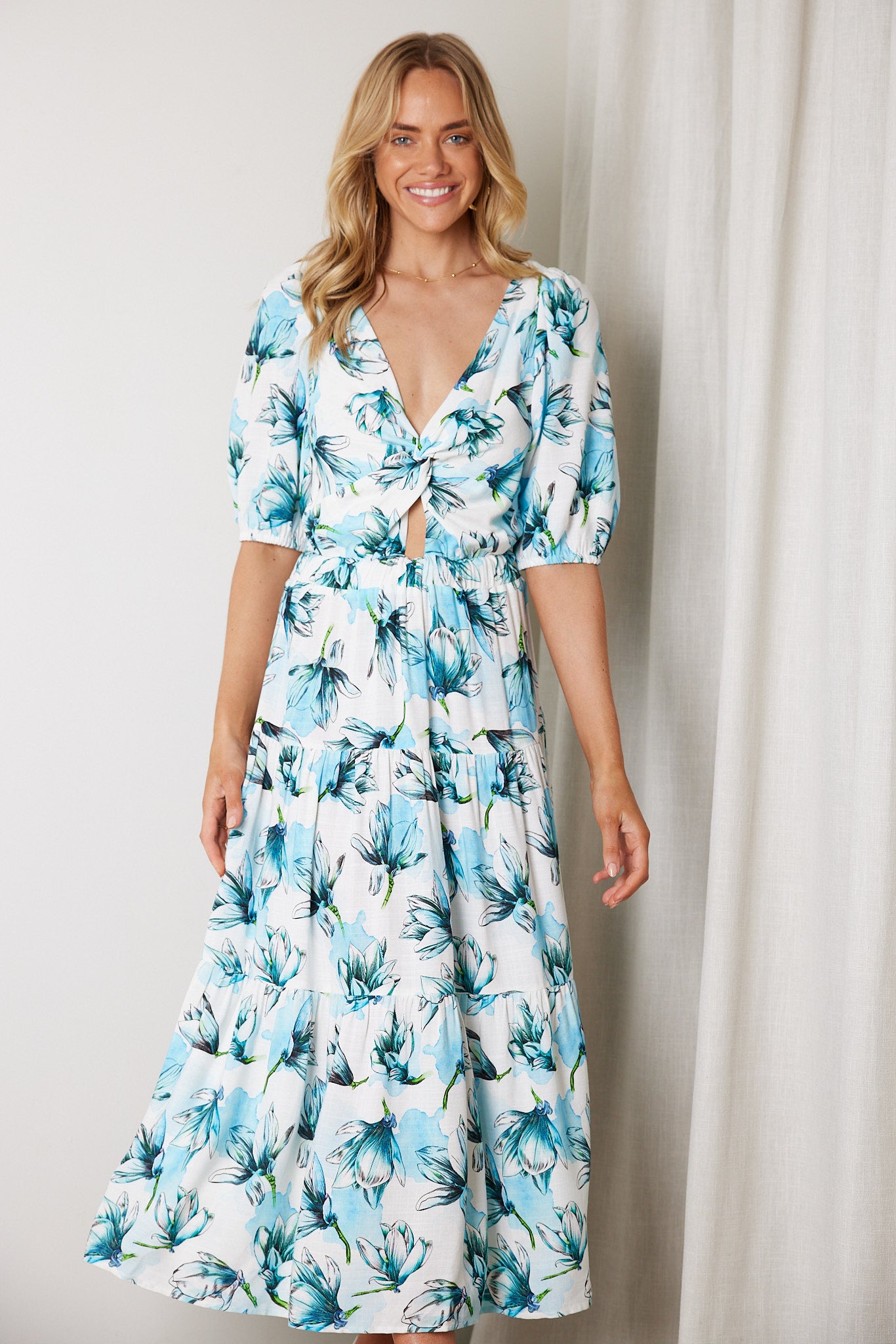 Woman wearing a white and blue floral midi dress with a front cut-out and ruched detailing, featuring elbow-length sleeves and a flowy skirt, standing indoors and smiling.