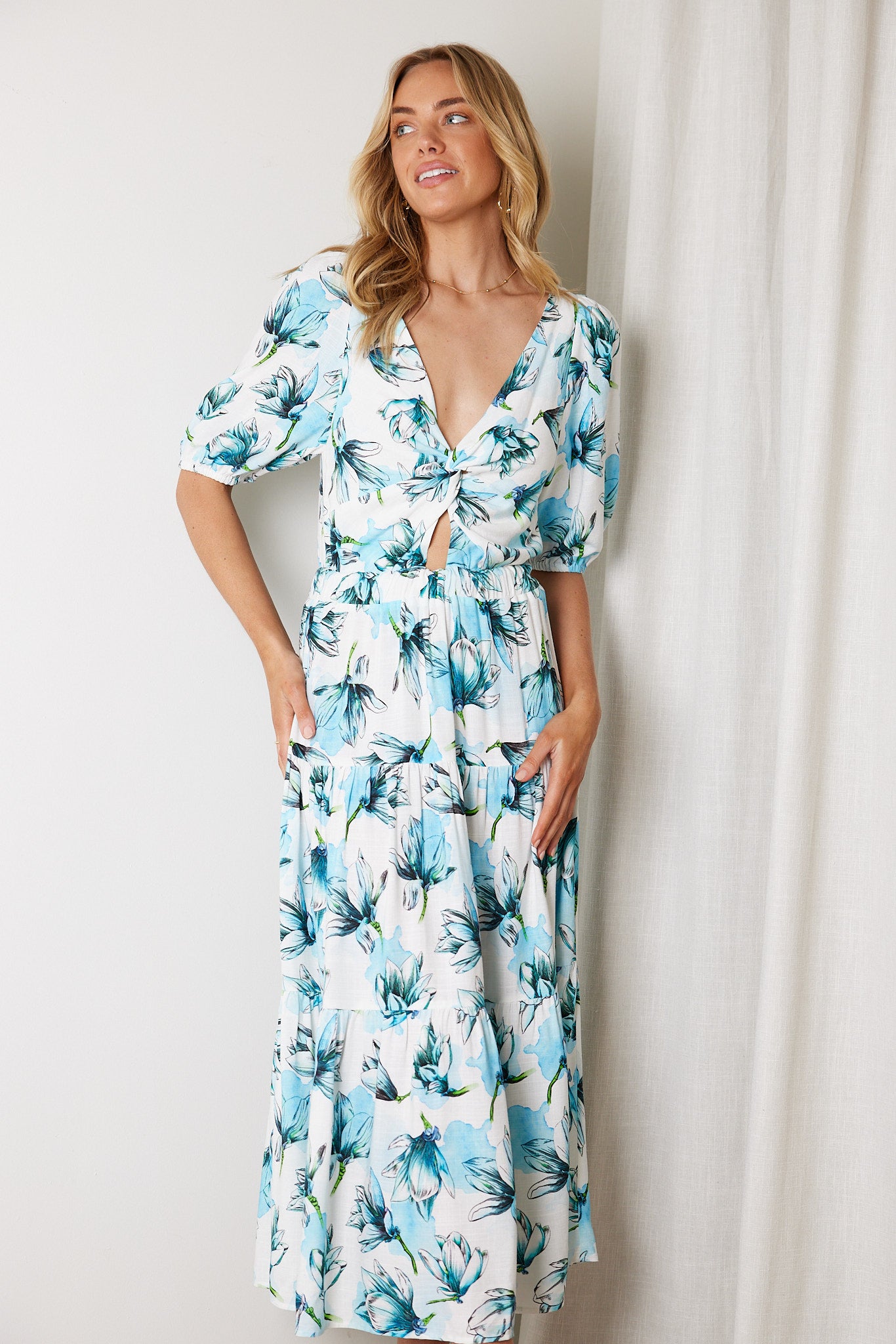 Woman wearing a white and blue floral midi dress with a front cut-out and ruched detailing, featuring elbow-length sleeves and a flowy skirt, standing indoors and smiling.