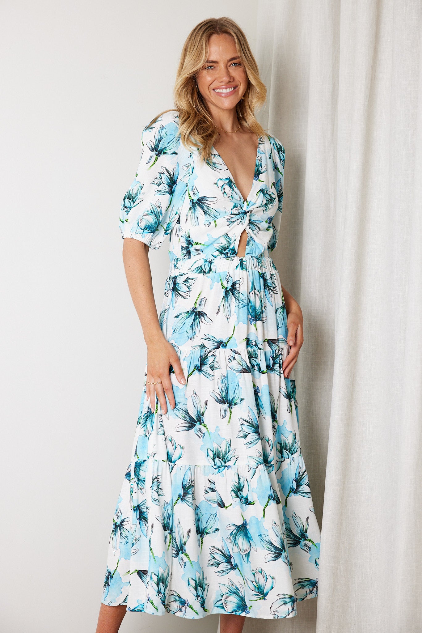 Woman wearing a white and blue floral midi dress with a front cut-out and ruched detailing, featuring elbow-length sleeves and a flowy skirt, standing indoors and smiling.