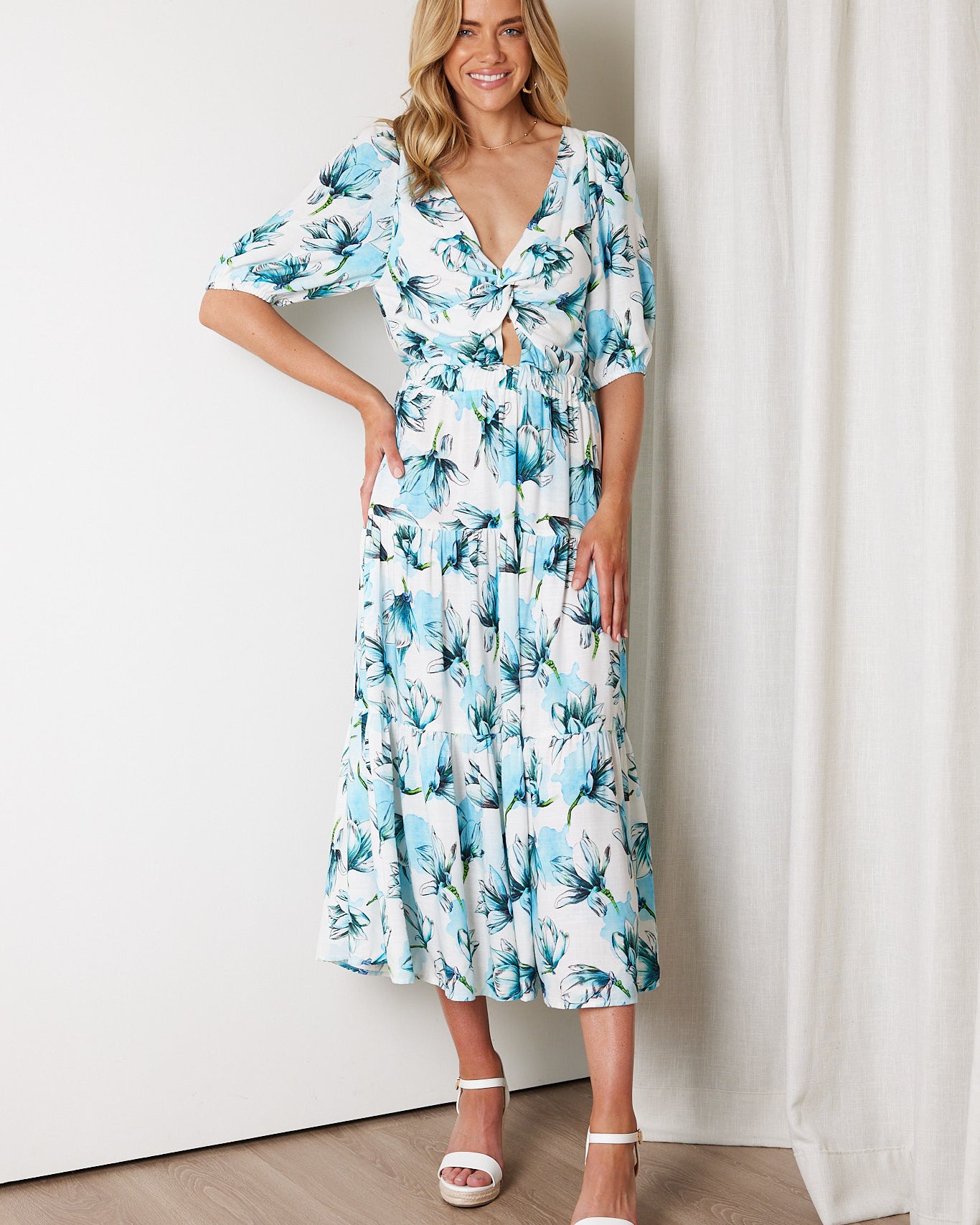 Woman wearing a white and blue floral midi dress with a front cut-out and ruched detailing, featuring elbow-length sleeves and a flowy skirt, standing indoors and smiling.