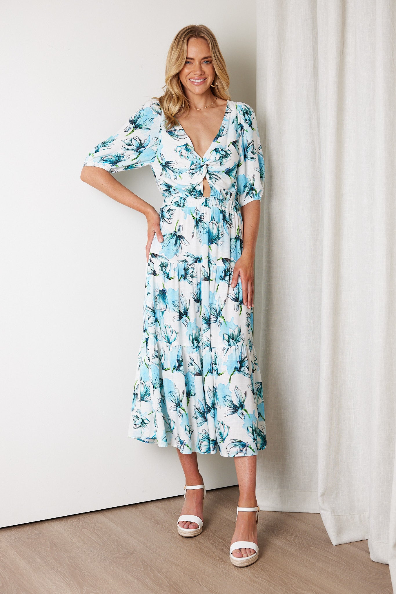 Woman wearing a white and blue floral midi dress with a front cut-out and ruched detailing, featuring elbow-length sleeves and a flowy skirt, standing indoors and smiling.